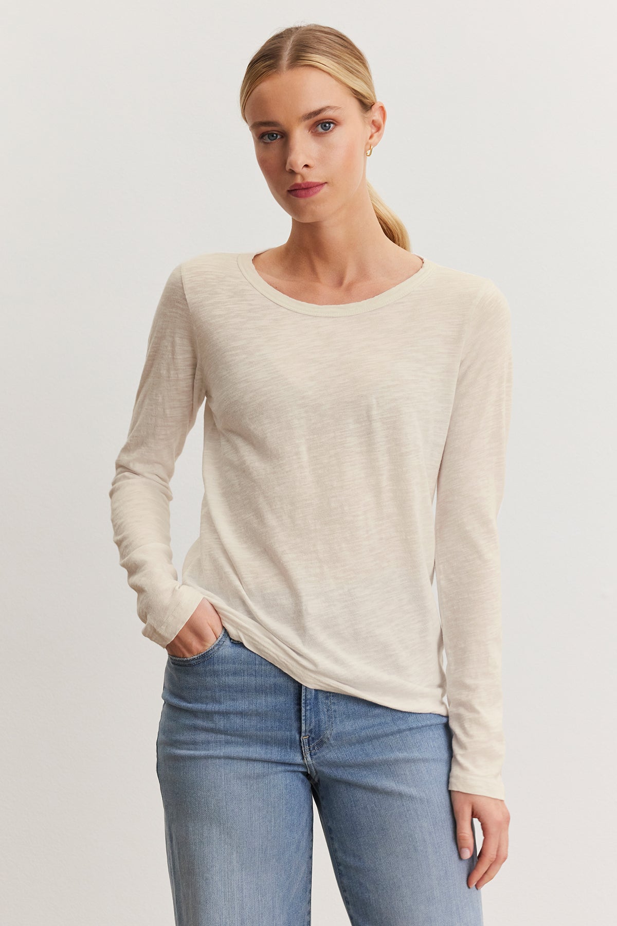   A person poses against a neutral background in blue jeans and the Velvet by Graham & Spencer LIZZIE TEE, a cream long-sleeve shirt with a classic crew neckline, with one hand in their pocket. 