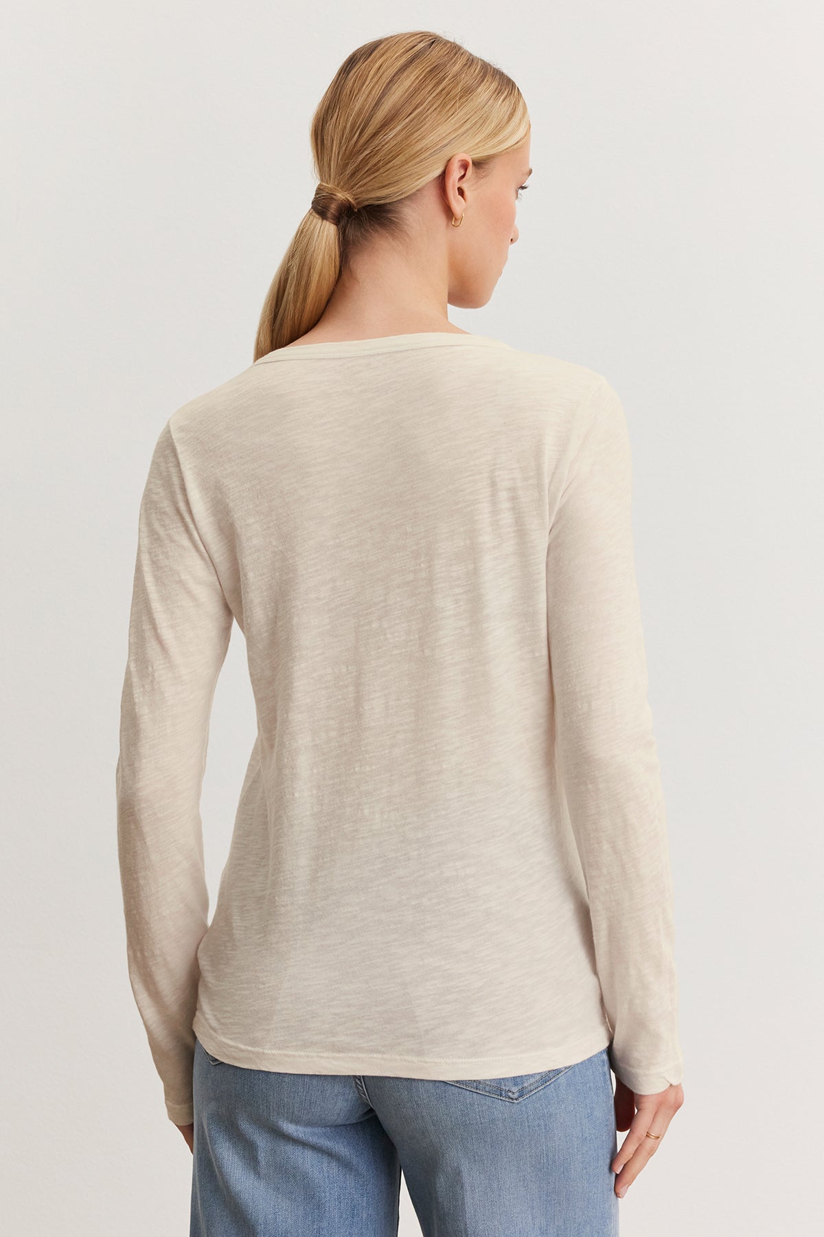   A person with long hair in a ponytail wears the LIZZIE TEE by Velvet by Graham & Spencer, featuring a beige textured cotton slub long-sleeve design, paired with blue jeans, facing away from the camera against a plain background. 