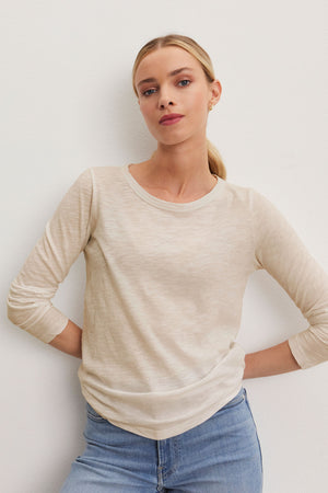 Against a plain background, someone with a ponytail is wearing the LIZZIE TEE, a long-sleeve beige top with a crew neckline from Velvet by Graham & Spencer, paired with jeans.