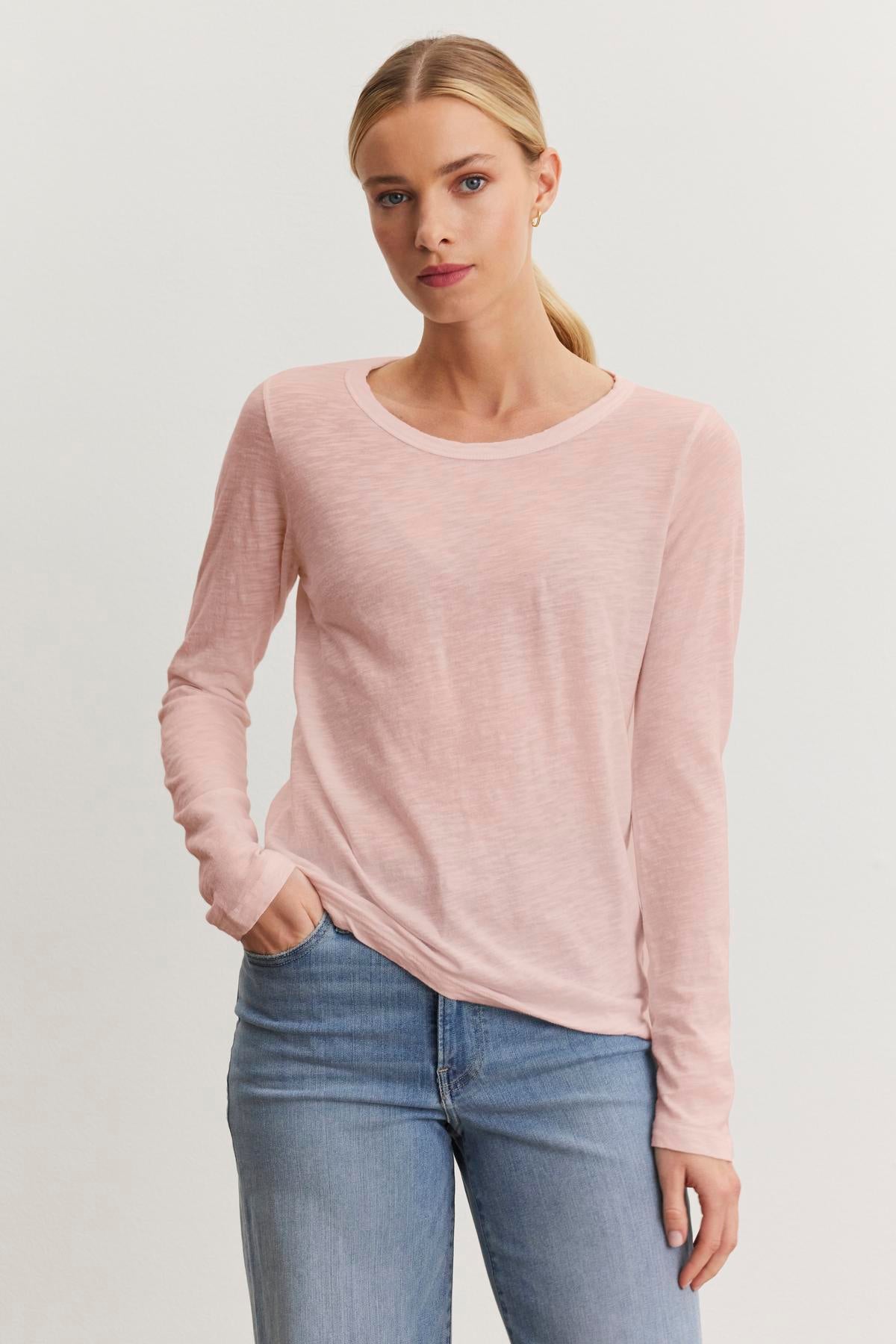 A person stands against a plain background, wearing the LIZZIE TEE in light pink by Velvet by Graham & Spencer, paired with blue jeans.-38301515219137