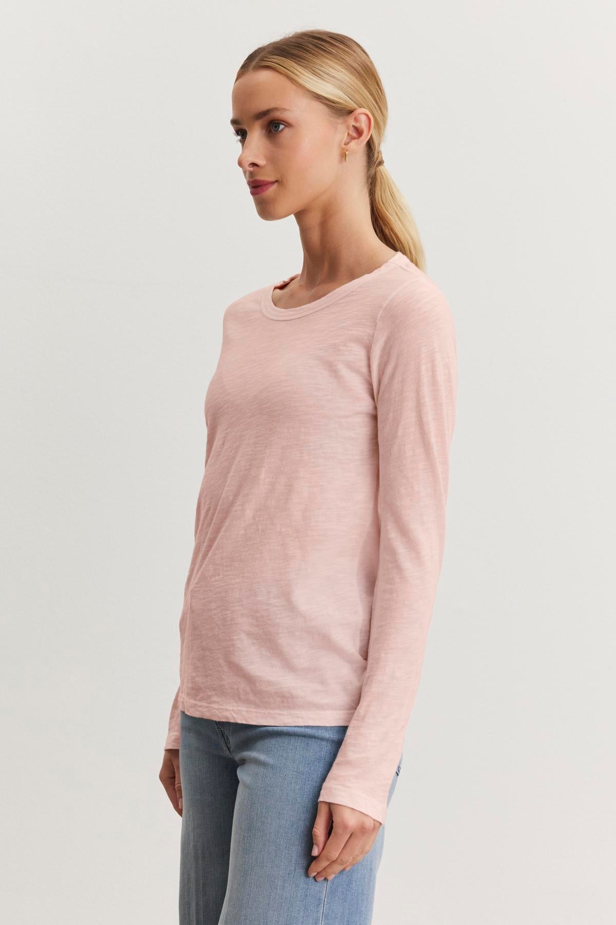   A person with a blond ponytail stands against a plain white background, wearing the LIZZIE TEE, a light pink cotton slub long sleeve top by Velvet by Graham & Spencer with a crew neckline, paired with blue jeans. 