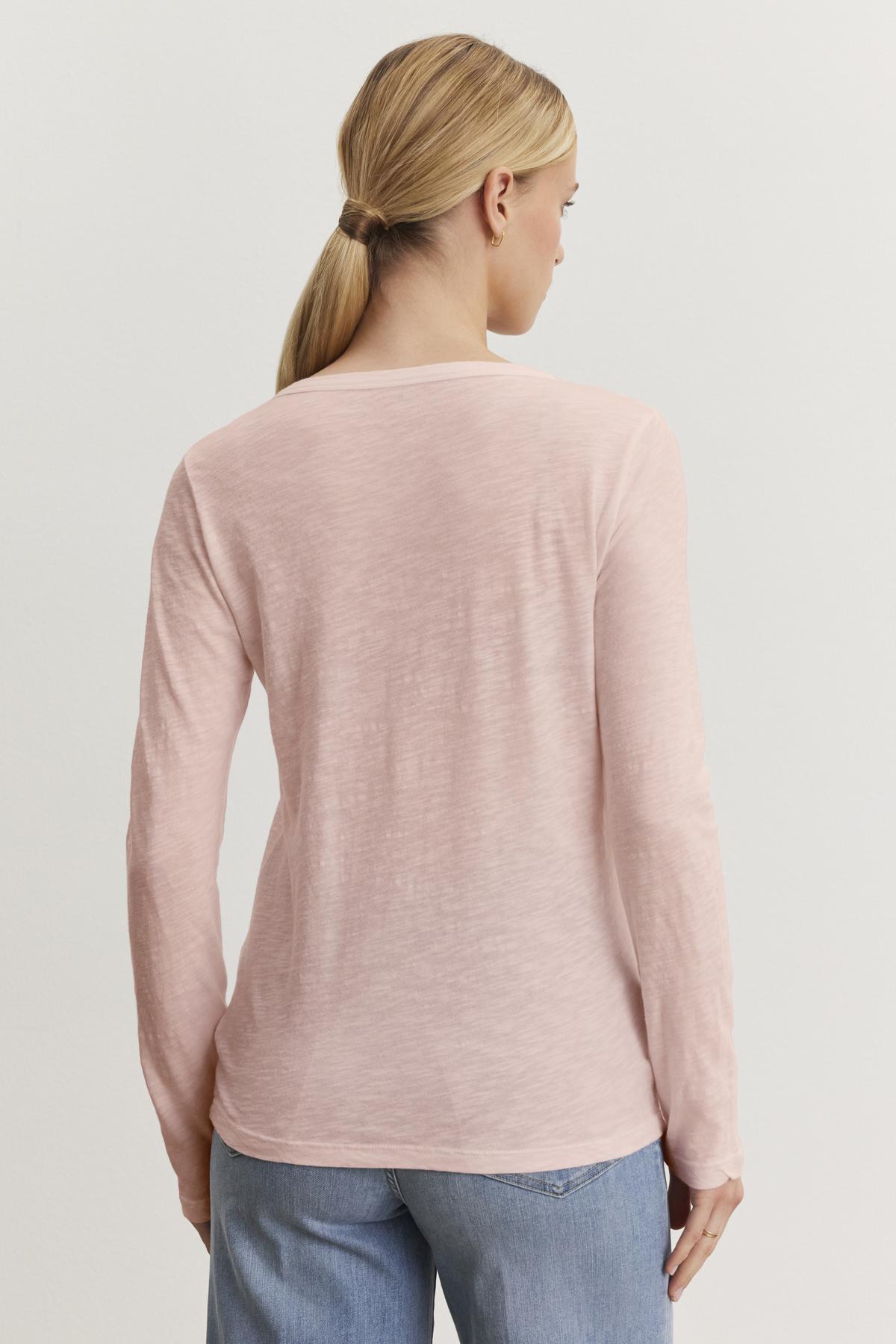   A person with long hair tied back is wearing the LIZZIE TEE, a light pink cotton slub long-sleeve top from Velvet by Graham & Spencer, paired with blue jeans, and is standing with their back to the camera against a plain background. 