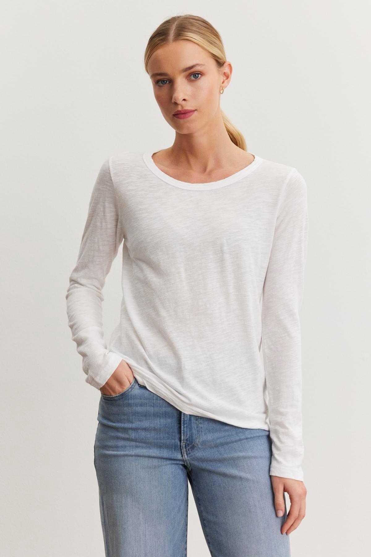   A person with blonde hair in a ponytail is wearing a white long-sleeve, classic crew neckline LIZZIE TEE by Velvet by Graham & Spencer and blue jeans, standing against a plain white background. 