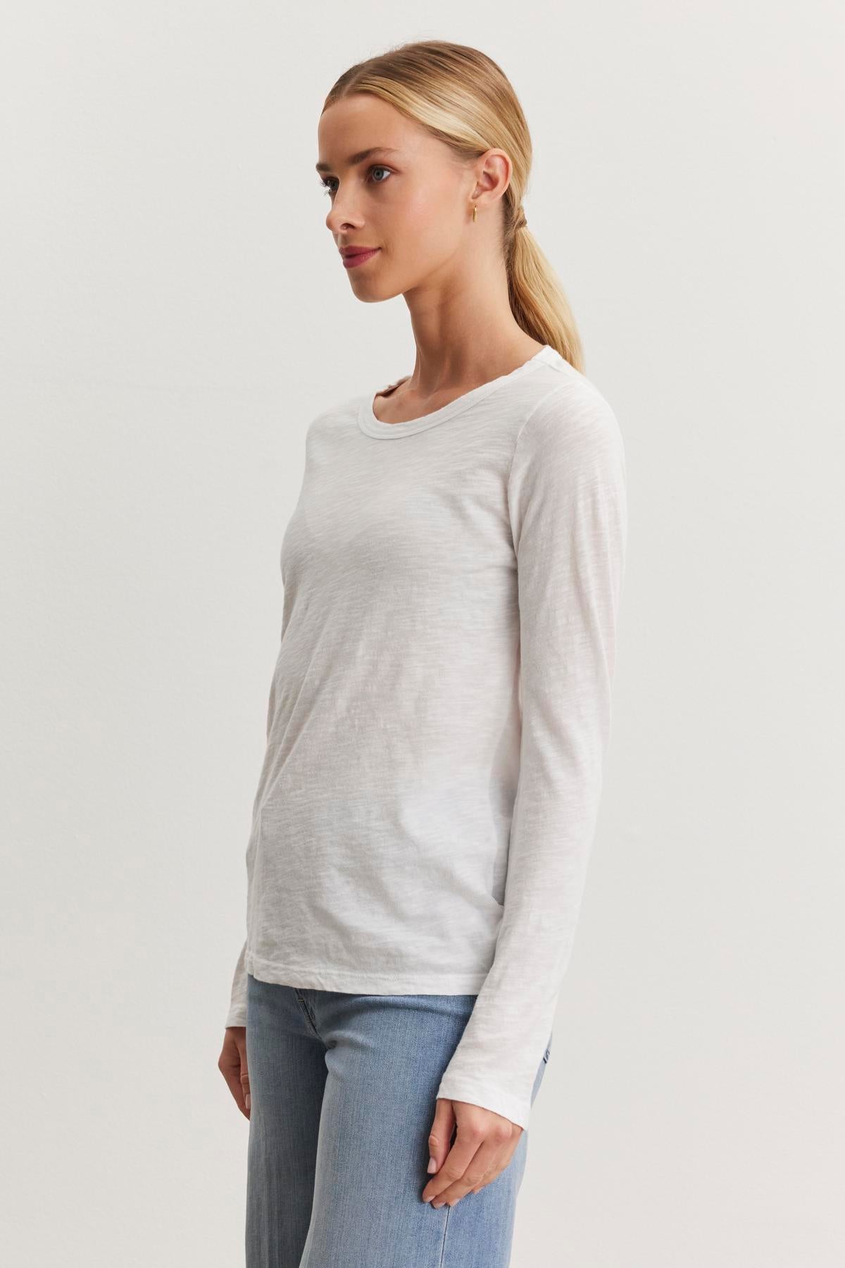   A person with blonde hair tied back in a ponytail is wearing the LIZZIE TEE by Velvet by Graham & Spencer and blue jeans, standing against a plain white background. 
