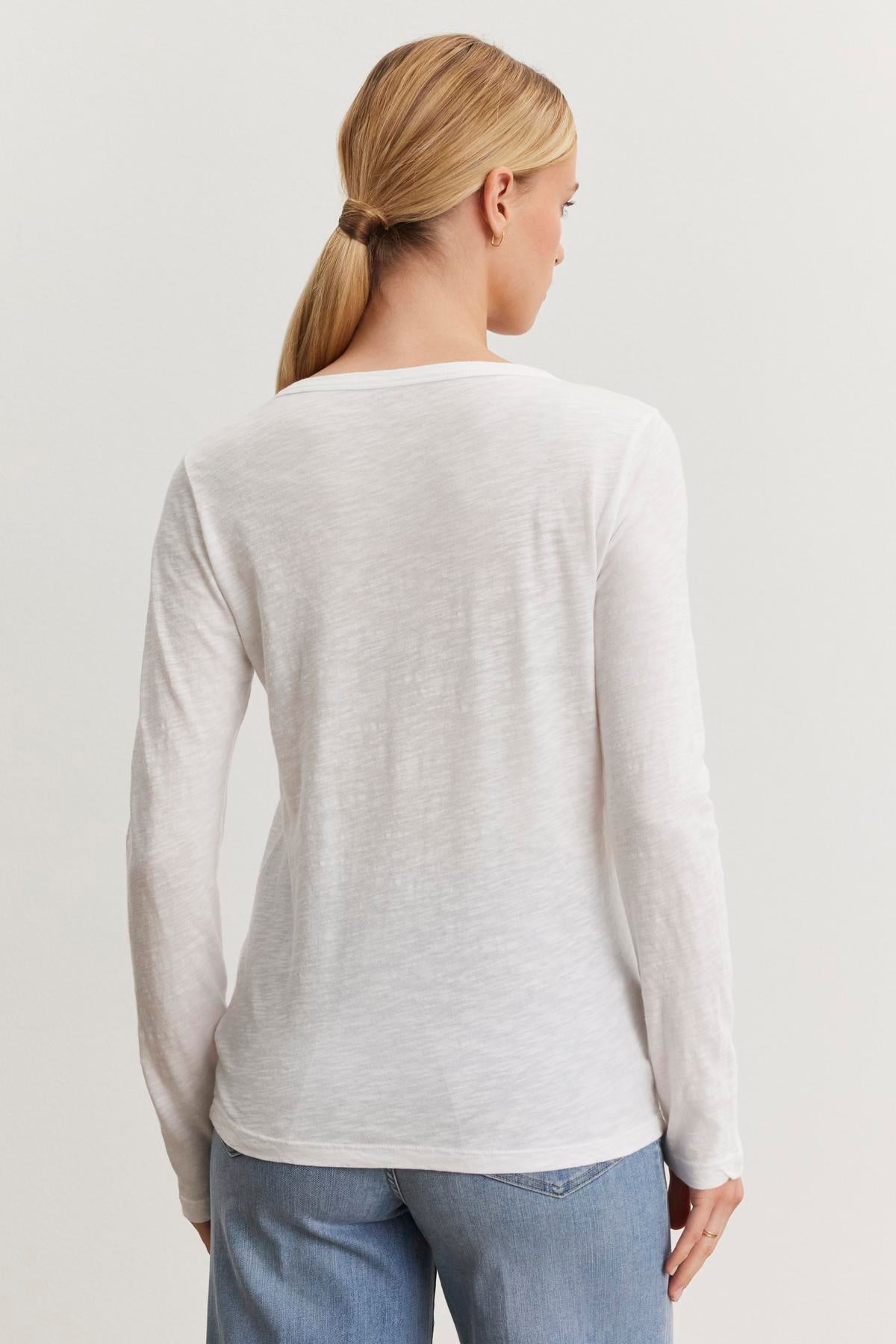   A person with blonde hair in a ponytail is shown from the back, wearing a long-sleeved white Velvet by Graham & Spencer LIZZIE TEE with a classic crew neckline and light blue jeans. 