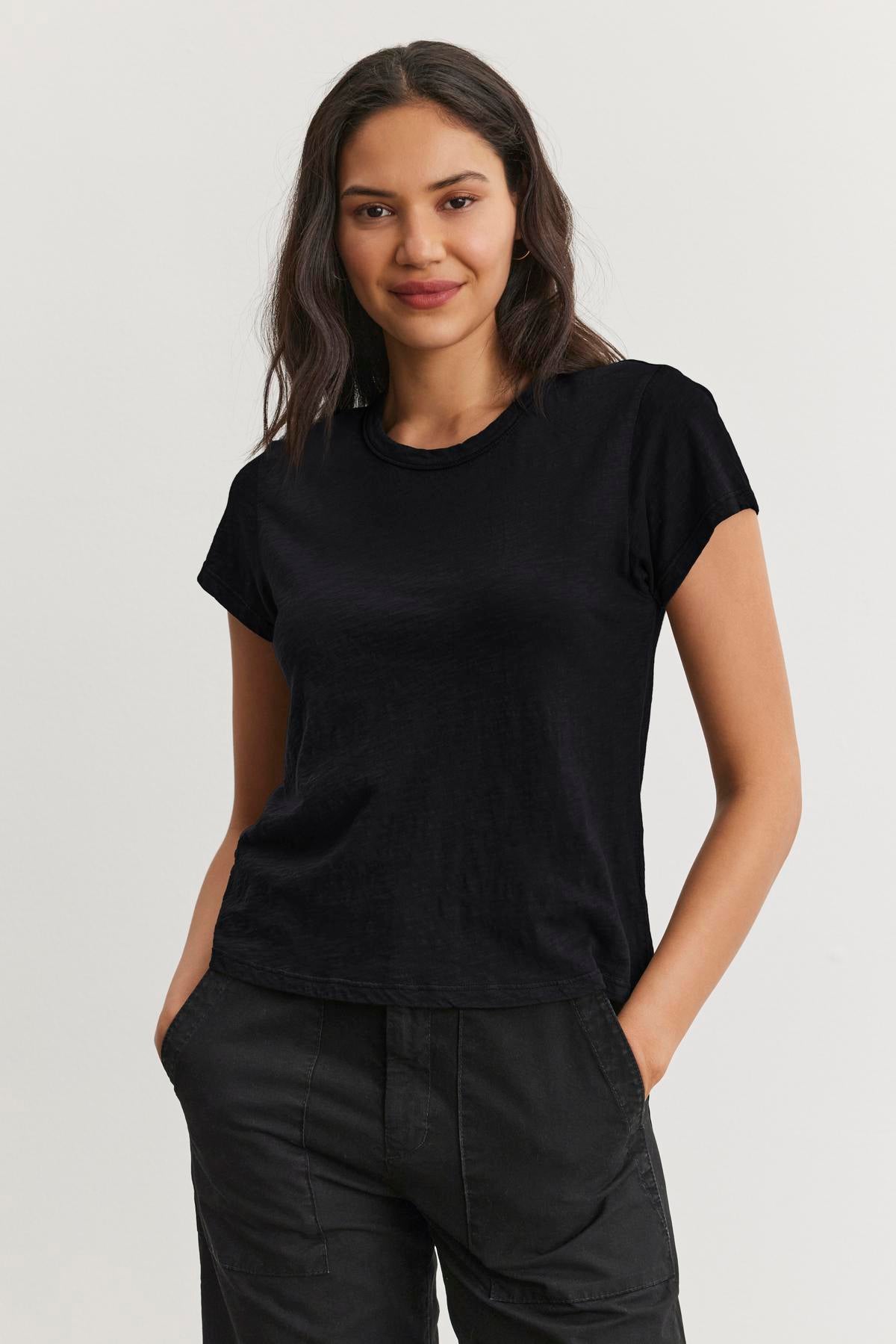   A person with long dark hair wears the MARIE TEE by Velvet by Graham & Spencer, a black cotton slub t-shirt with a boxy fit, along with dark pants, against a plain background. 