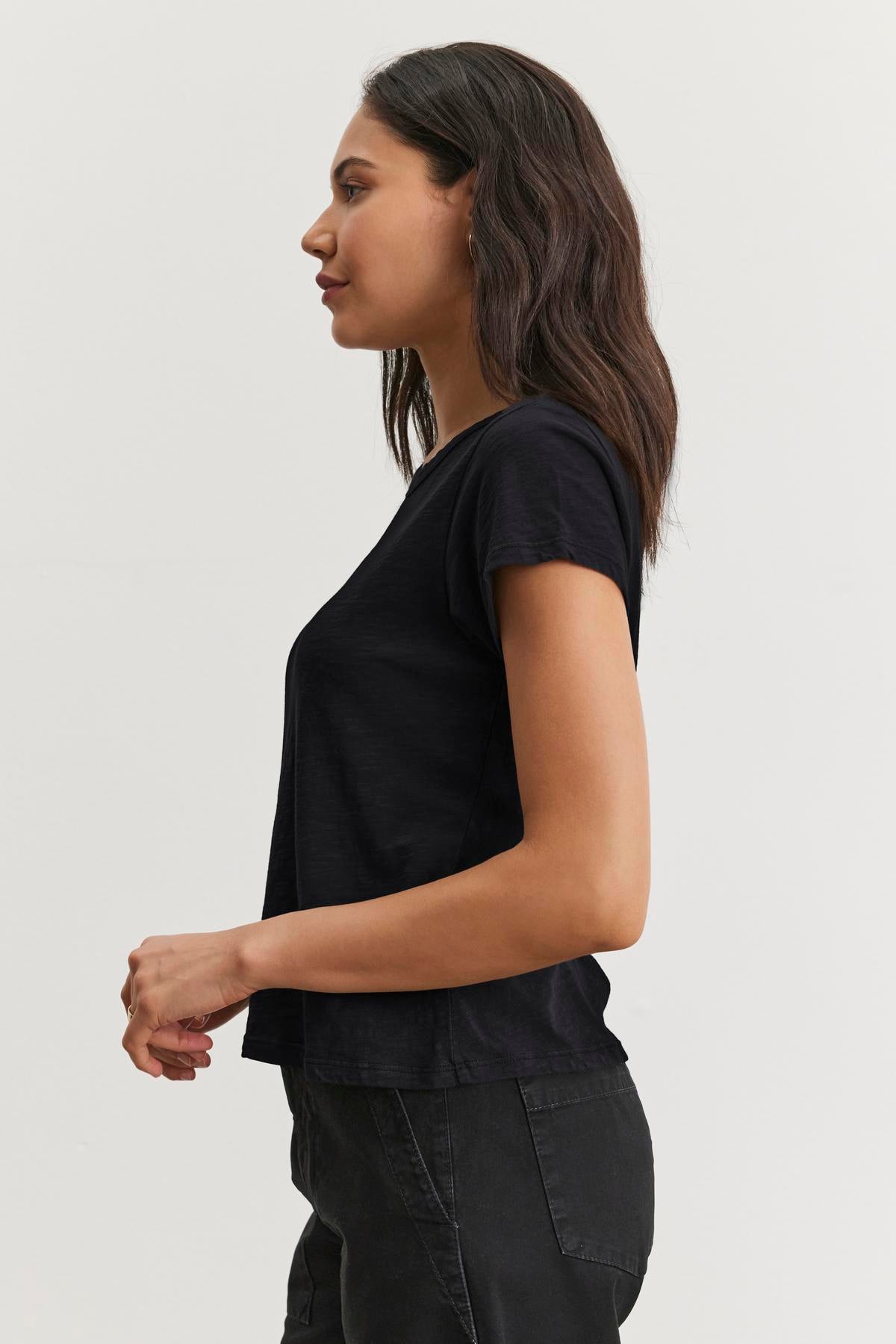   A woman wearing the MARIE TEE from Velvet by Graham & Spencer with pants stands in profile against a plain background, perfectly embodying a casual occasion. 