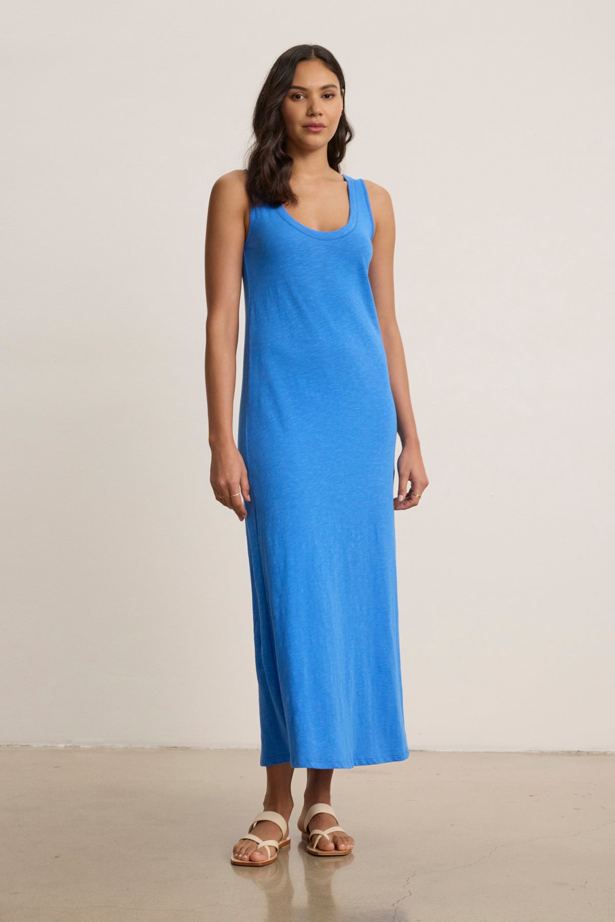 A woman wears the CAMILA DRESS by Velvet by Graham & Spencer, a sleeveless, ankle-length blue maxi dress paired with white sandals against a plain background—ideal for everyday wear.