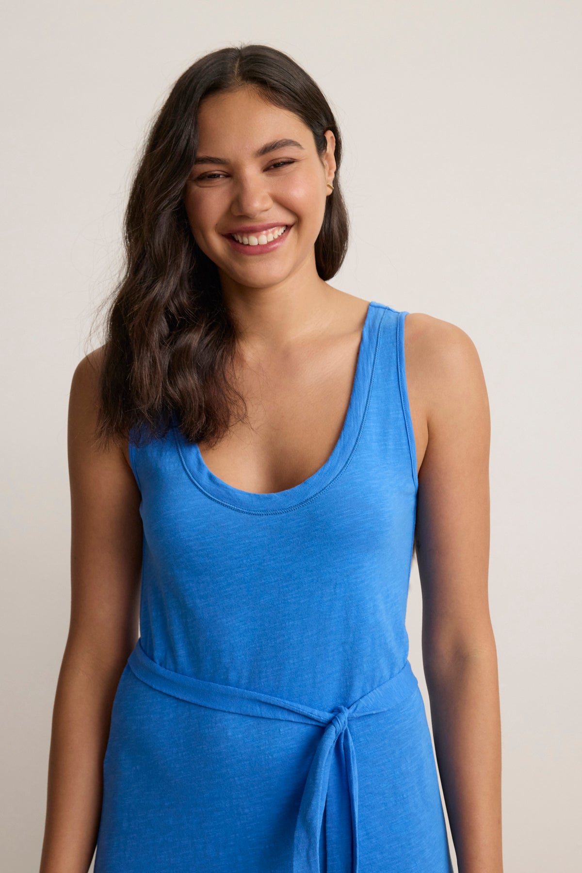 A woman with long dark hair, wearing the CAMILA DRESS by Velvet by Graham & Spencer—a sleeveless blue textured cotton slub maxi dress with a waist tie—stands smiling against a neutral background.-Hover Image