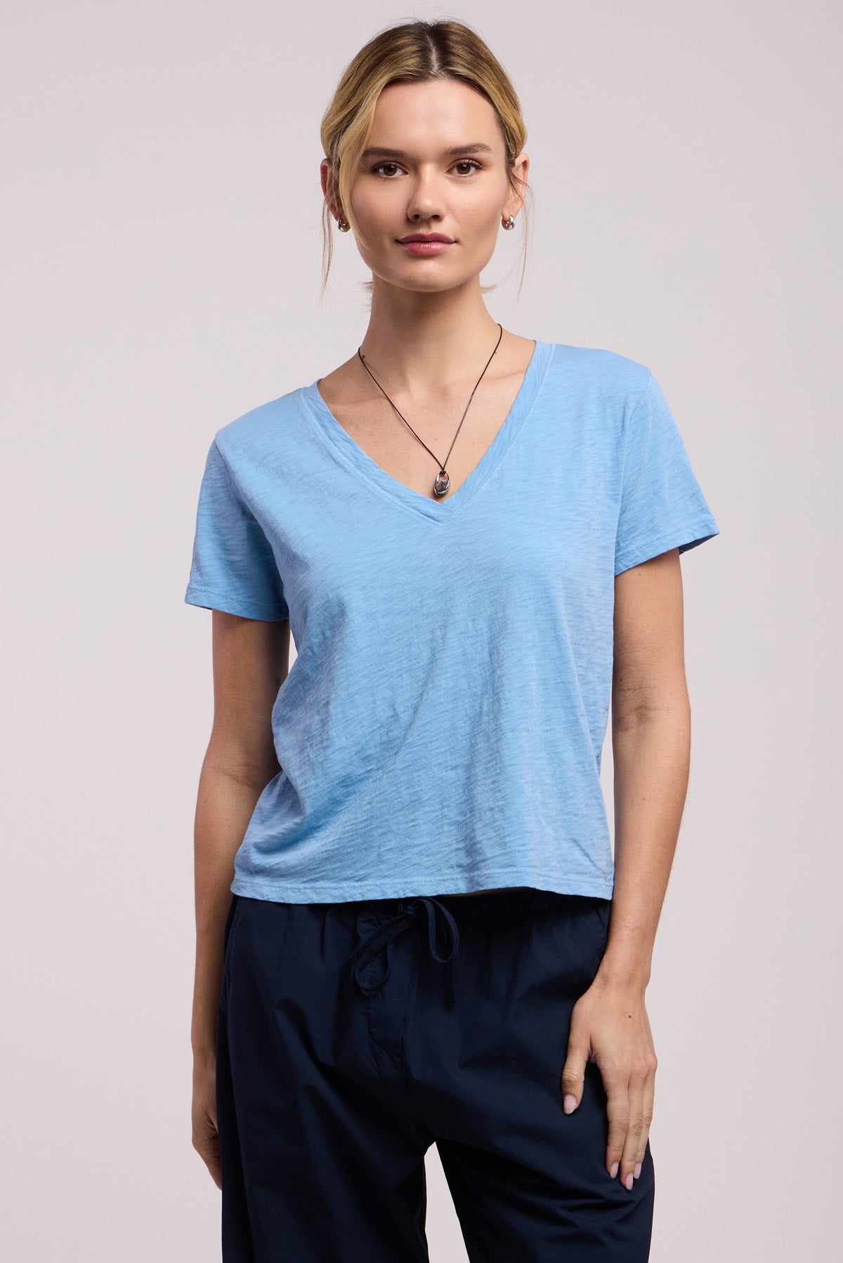 Wearing the ELISHA TEE by Velvet by Graham & Spencer in light blue cotton slub with a V-neck, paired with dark blue pants and a necklace, someone stands against a plain light background—a true wardrobe staple.-41645010944193