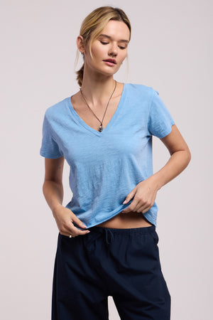 A woman stands against a plain background, looking slightly downward and holding the hem of the ELISHA TEE by Velvet by Graham & Spencer—a light blue cotton slub V-neck—paired with dark pants.