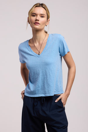 Person wears the ELISHA TEE by Velvet by Graham & Spencer—a light blue cotton slub V-neck—paired with dark drawstring pants, standing against a plain background with one hand in their pocket and a neutral expression.