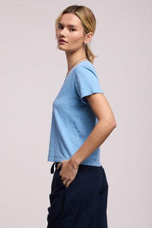 A person stands in a relaxed pose, facing sideways, wearing the ELISHA TEE by Velvet by Graham & Spencer in light blue paired with dark pants, set against a neutral background.