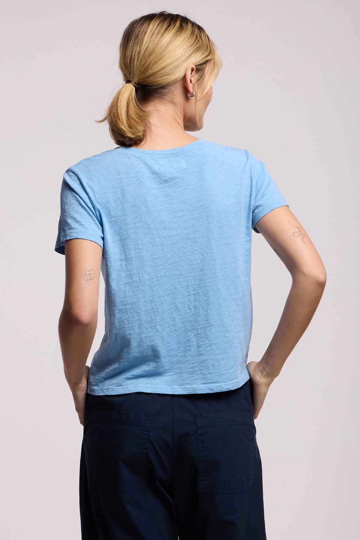 A person with blonde hair in a ponytail is shown from the back, wearing the Velvet by Graham & Spencer ELISHA TEE in light blue and dark blue pants, standing against a plain background.-41645011206337