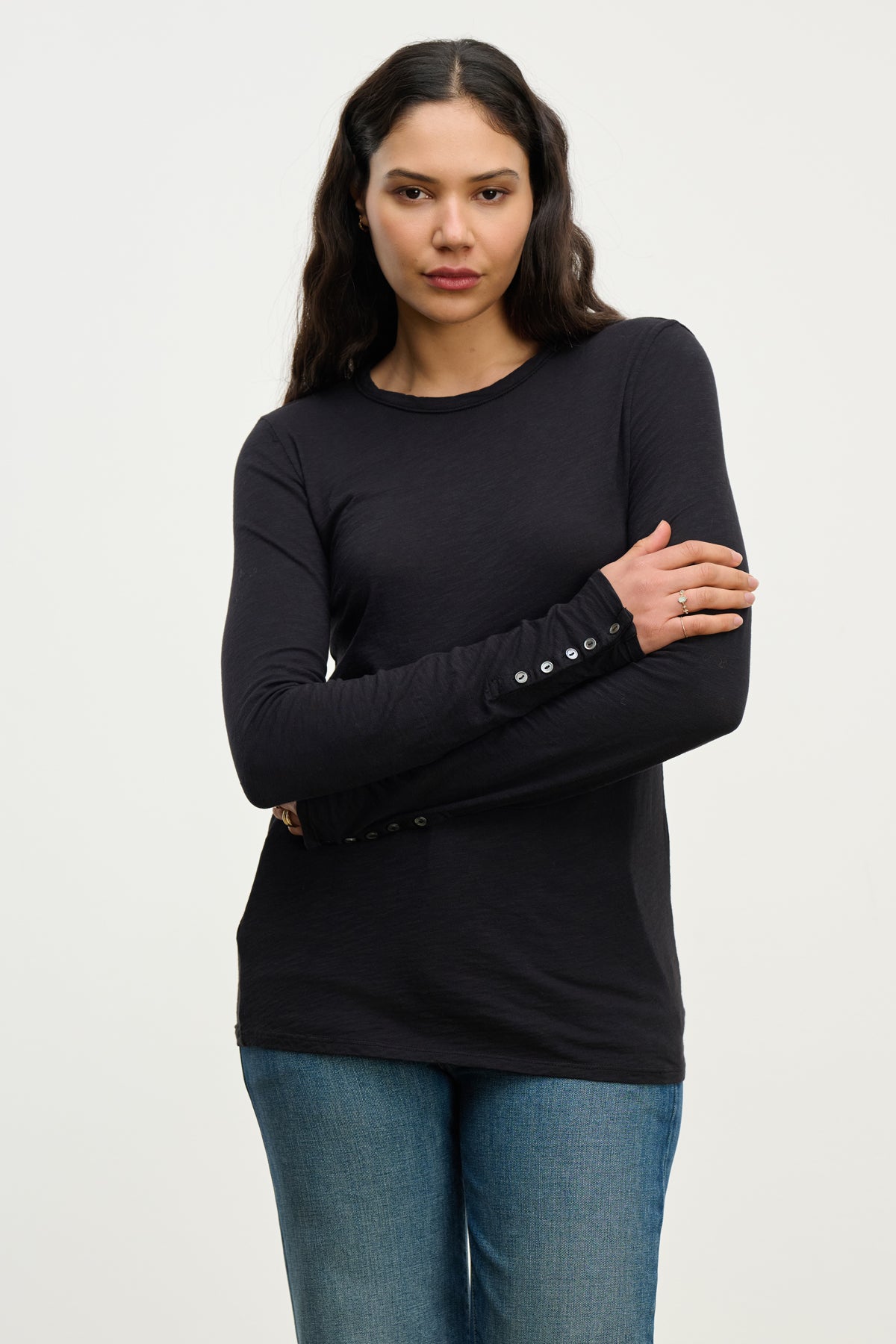 A woman with long dark hair models the KELSEY TEE by Velvet by Graham & Spencer—a black, long-length button sleeve top—paired with blue jeans, standing against a white background.-39815225835713