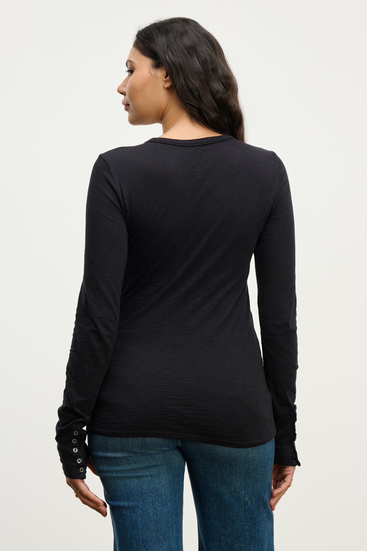 A woman with long dark hair is seen from the back, wearing the KELSEY TEE by Velvet by Graham & Spencer and blue jeans, standing against a plain light background.-39815225770177