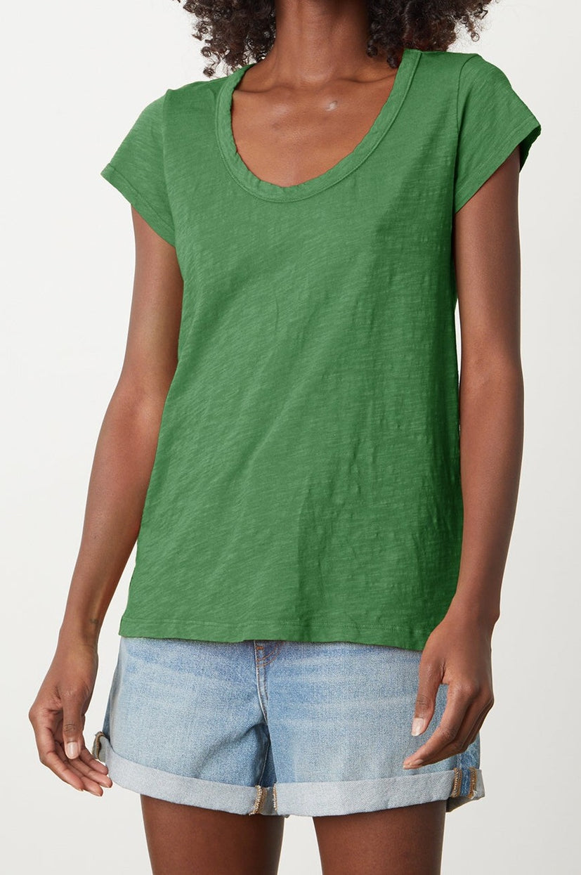 A person wearing a green KIRA TEE from Velvet by Graham & Spencer made from cotton slub fabric and blue denim shorts. The scoop neckline adds a casual touch to the everyday wear. The person is standing against a white background.