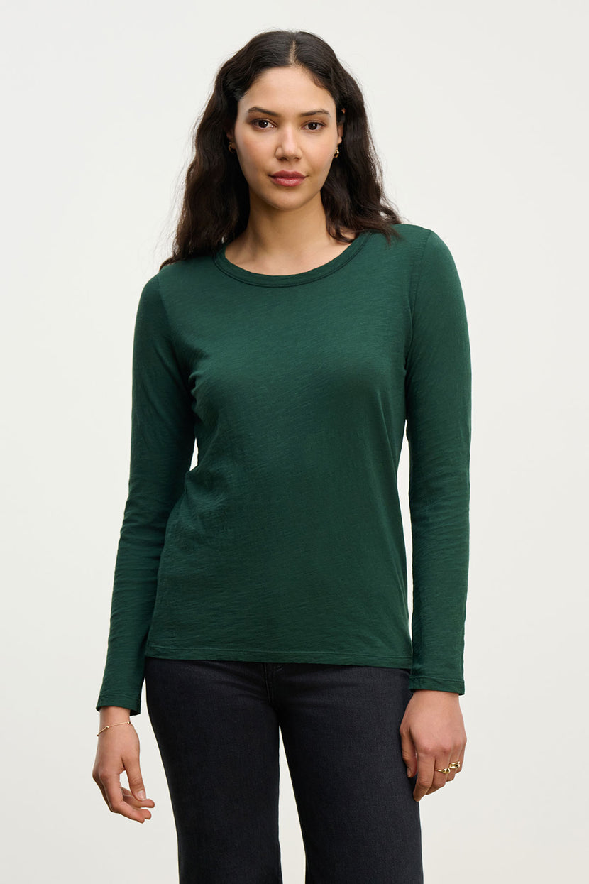 A woman with long dark hair wears the LIZZIE TEE by Velvet by Graham & Spencer in dark green, styled with black pants, standing against a plain light background.