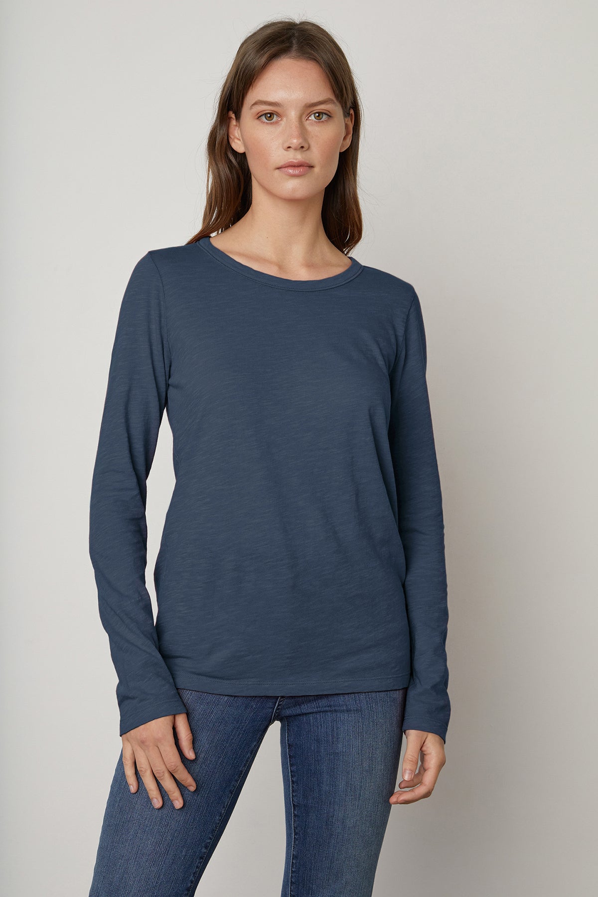   A person with long brown hair wearing a blue Velvet by Graham & Spencer LIZZIE TEE and blue jeans stands against a gray background. 