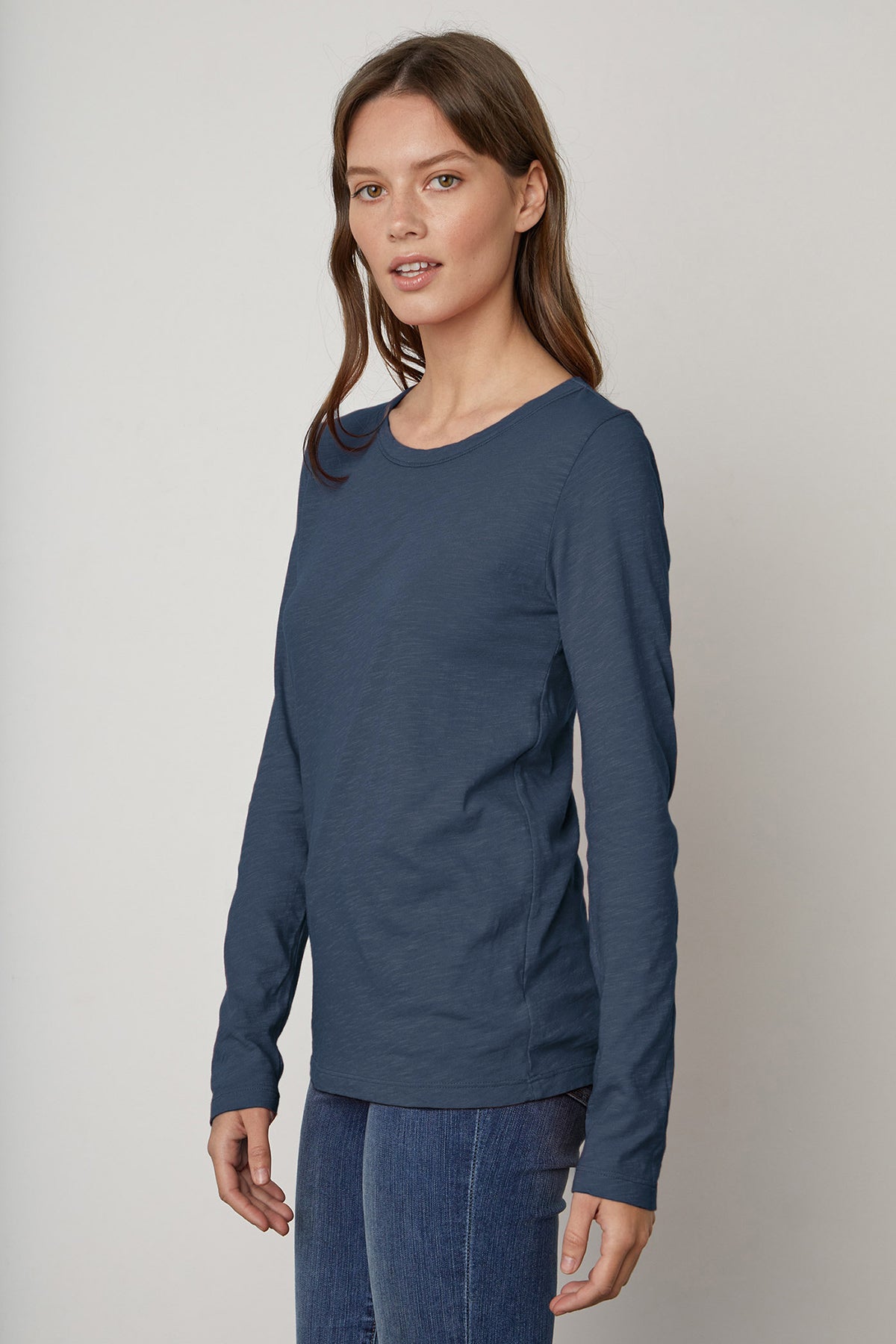   A woman with brown hair stands wearing the LIZZIE TEE, a long-sleeve, dark blue shirt with a classic crew neckline from Velvet by Graham & Spencer, paired with blue jeans. She is looking slightly to the side with a neutral expression. 