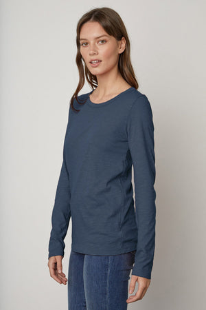 A woman with brown hair stands wearing the LIZZIE TEE, a long-sleeve, dark blue shirt with a classic crew neckline from Velvet by Graham & Spencer, paired with blue jeans. She is looking slightly to the side with a neutral expression.