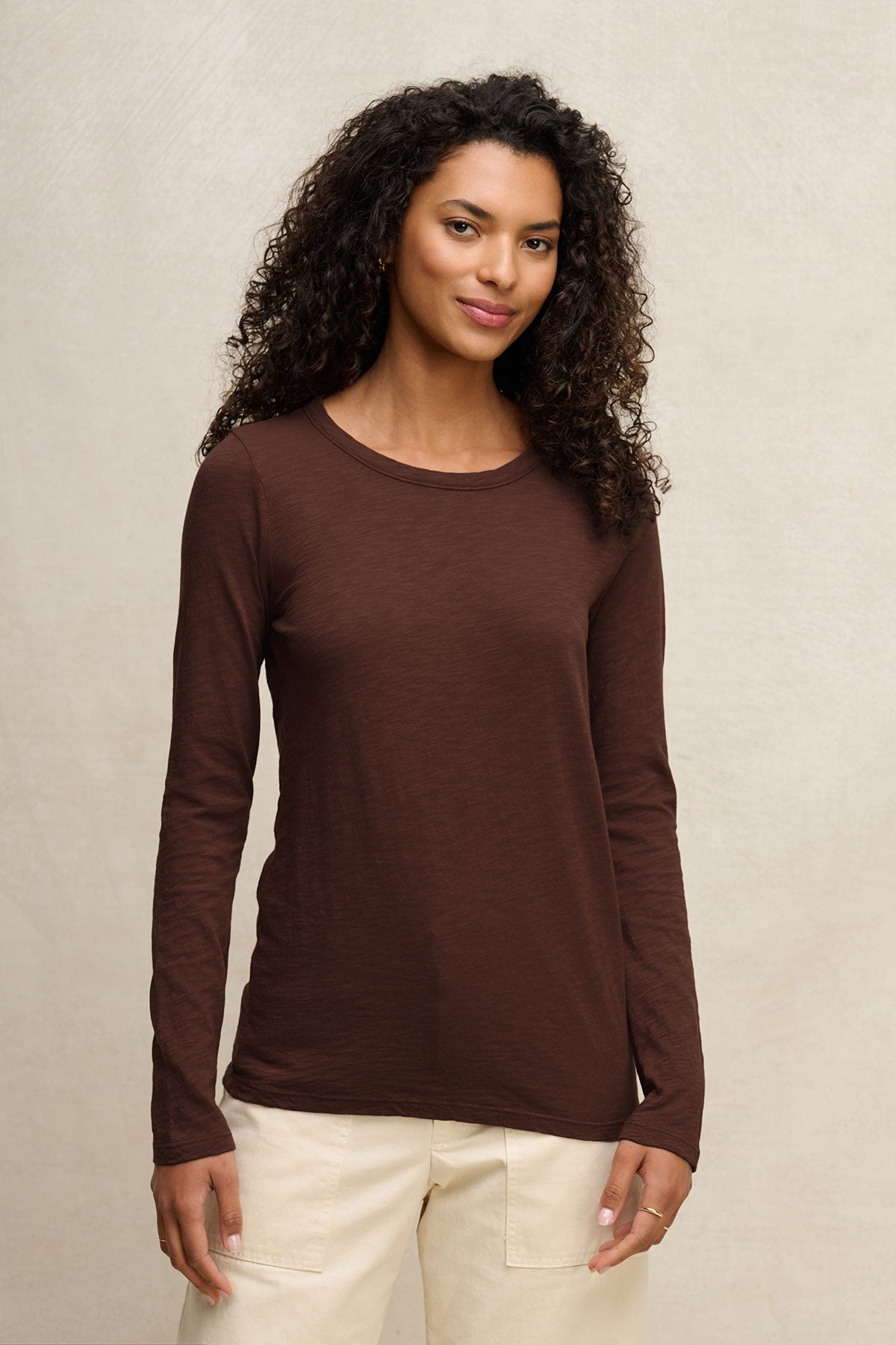 A woman with curly hair wears the Velvet by Graham & Spencer LIZZIE TEE with long sleeves and a crew neckline, paired with light-colored pants, standing against a neutral background.-40416960872641