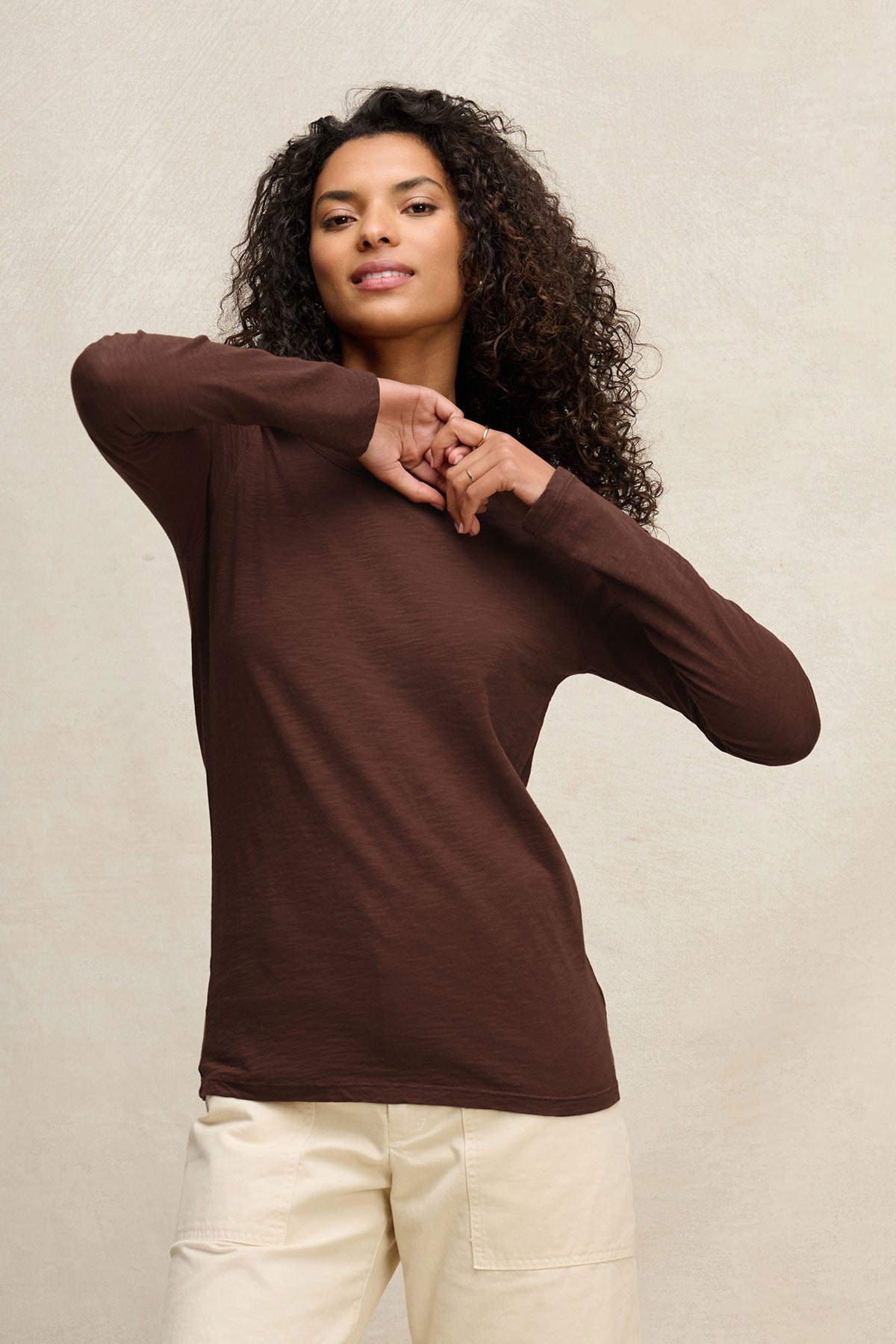 A woman poses against a neutral background wearing the Velvet by Graham & Spencer LIZZIE TEE—a long sleeve, crew neck brown textured cotton slub top—paired with light pants, her arms bent and hands near her shoulders.-40416960905409