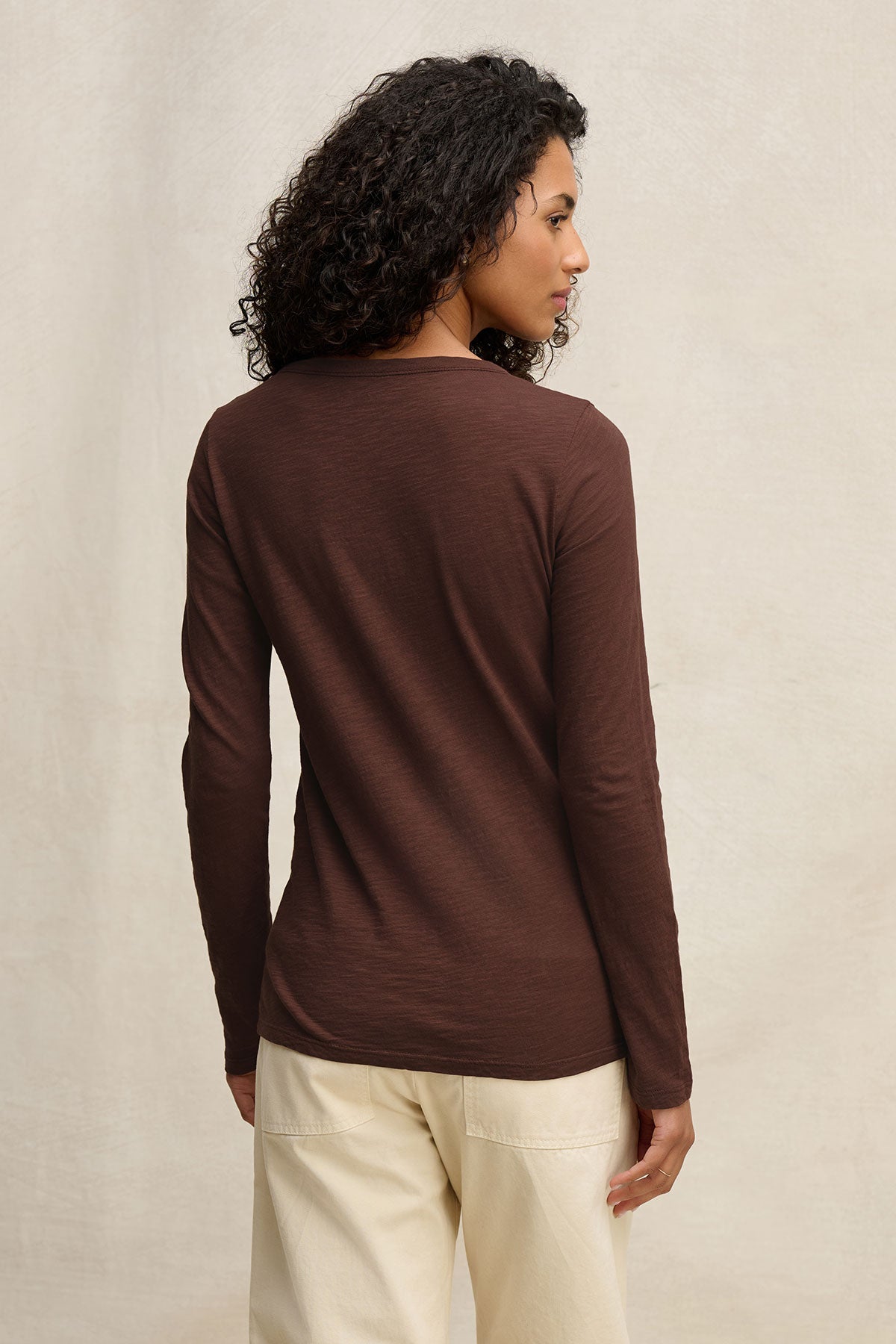 A person with curly hair wears the Velvet by Graham & Spencer LIZZIE TEE, a long sleeve crew neck top, paired with cream pants, standing facing away against a neutral background.-40416960970945