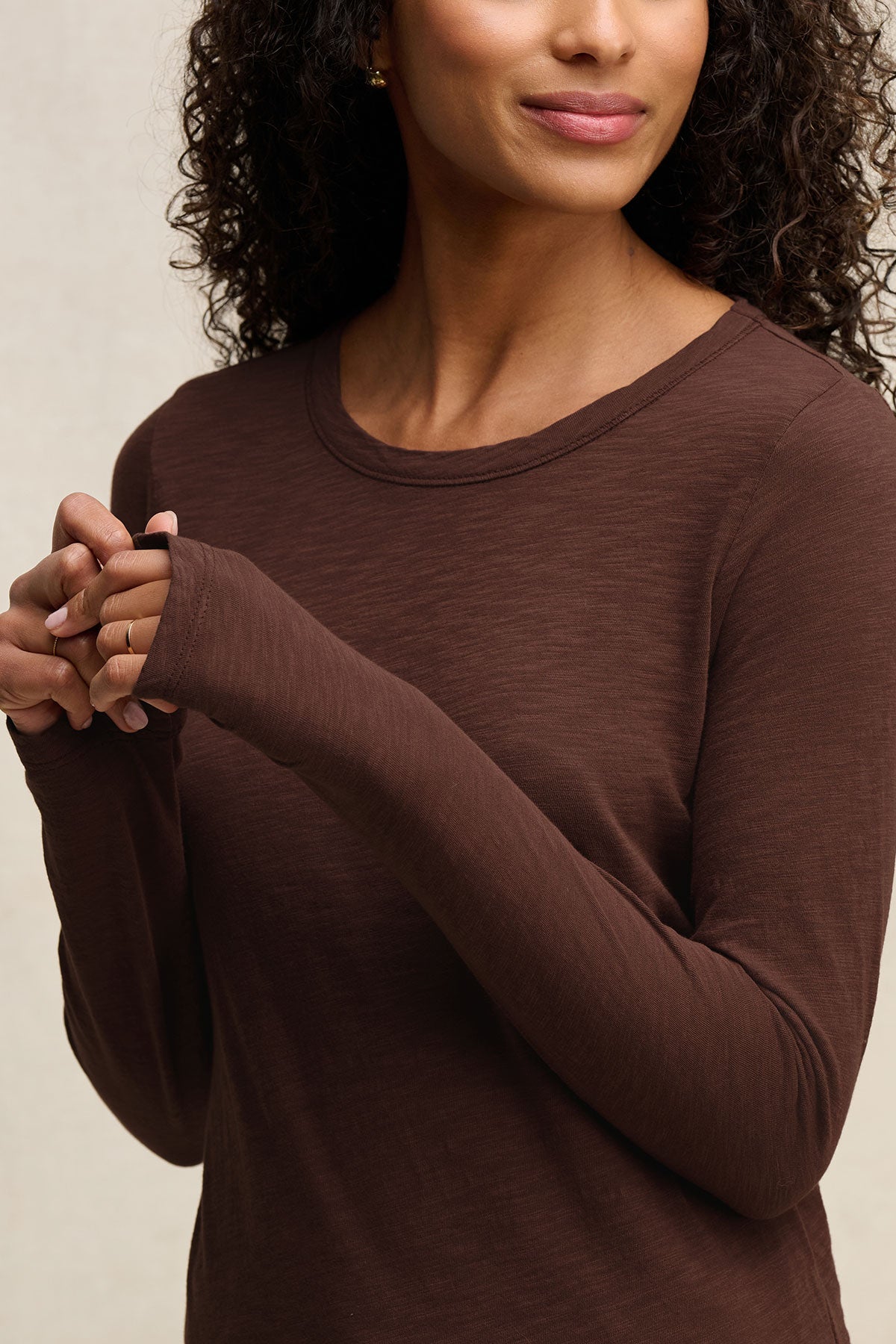 A woman wears the LIZZIE TEE by Velvet by Graham & Spencer, featuring long sleeves with thumbholes, a crew neckline, and textured cotton slub; she stands with hands clasped, upper body and part of her face visible.-40416960938177