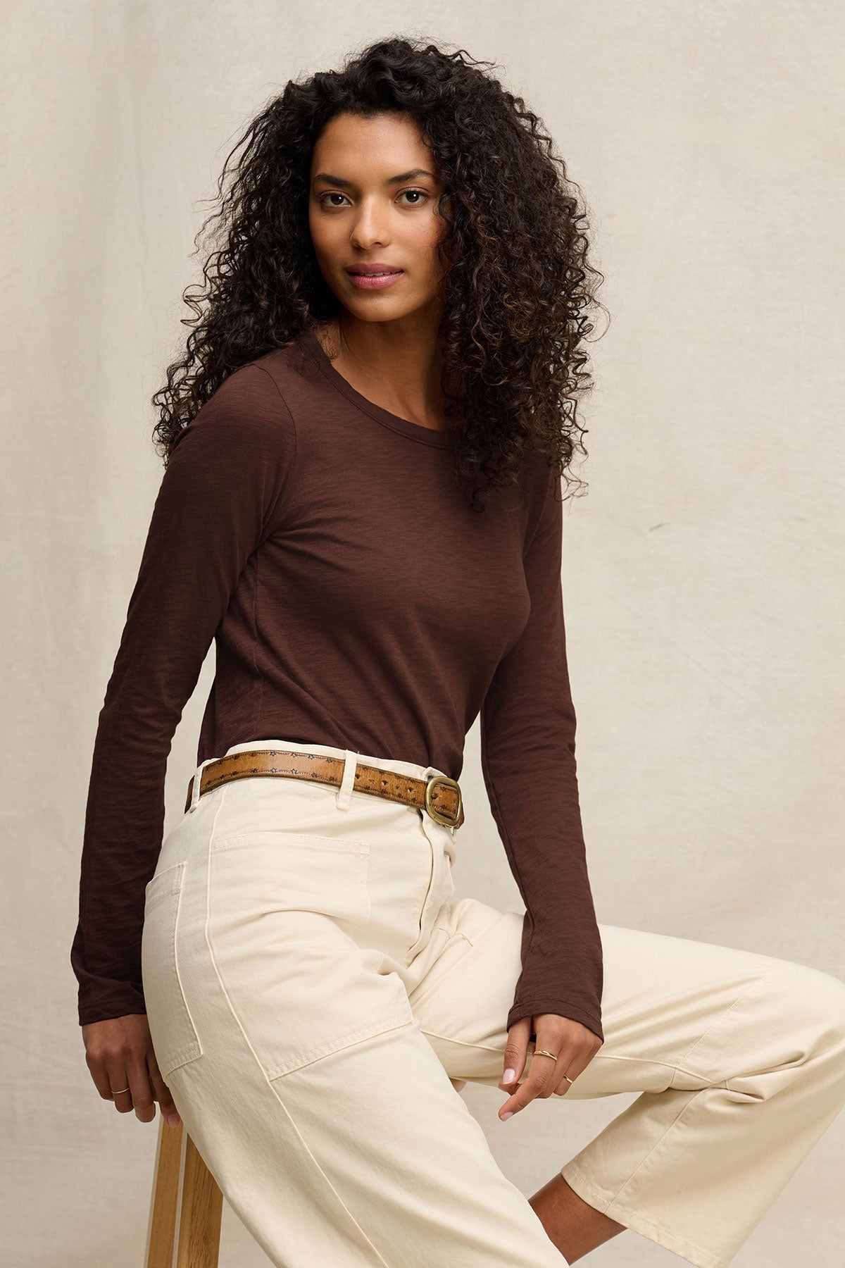 A woman with curly hair wears the LIZZIE TEE by Velvet by Graham & Spencer— a brown long sleeve crewneck—paired with beige pants and a brown belt as she sits on a stool against a light background.-40416961003713