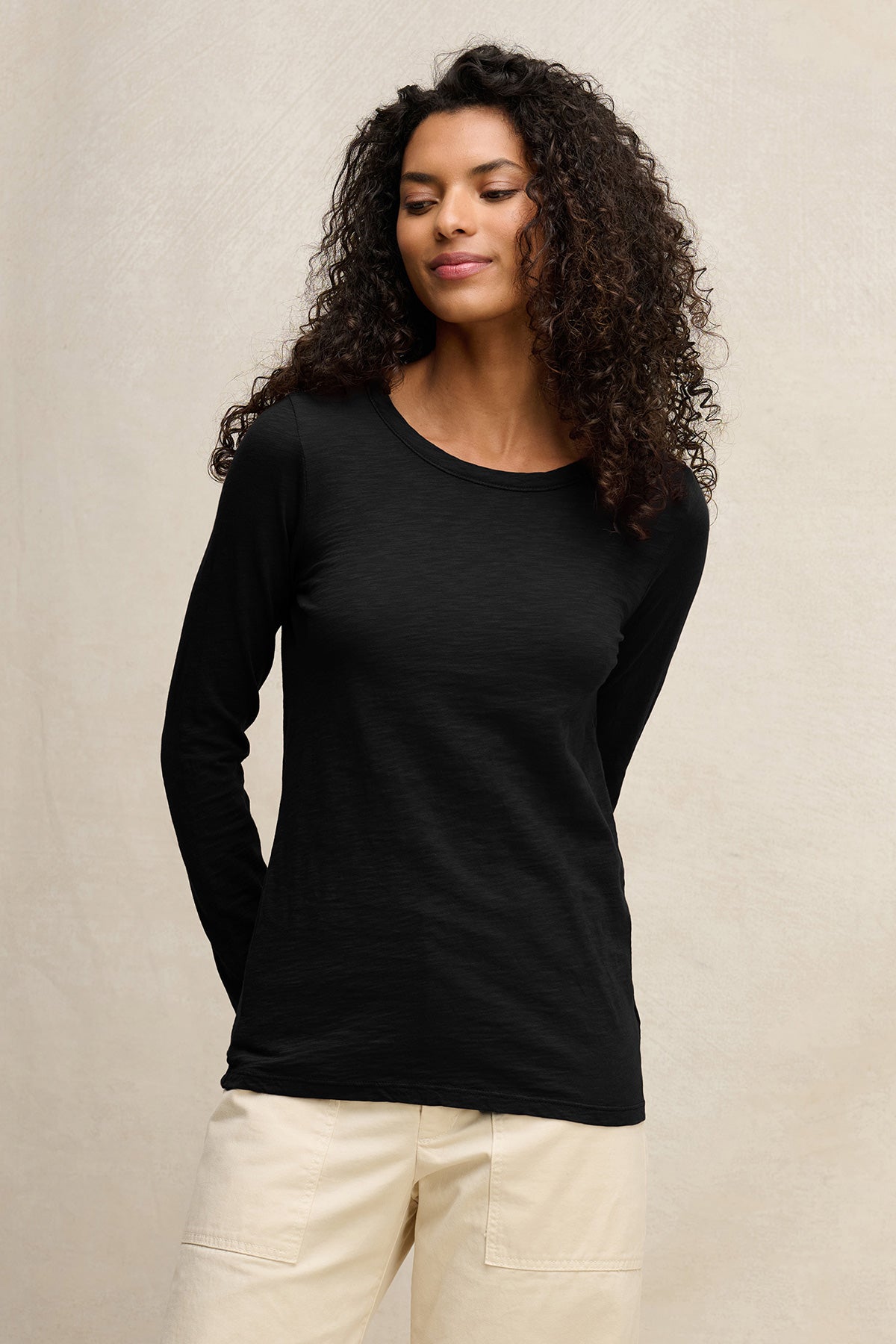 A woman with curly hair wears the LIZZIE TEE by Velvet by Graham & Spencer, a black cotton slub crewneck top, paired with light pants against a simple, light background.-40305805263041