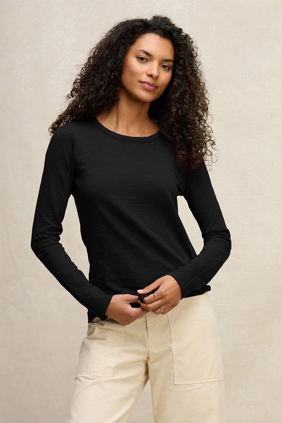 A woman with curly hair wears the LIZZIE TEE by Velvet by Graham & Spencer, a black long-sleeve cotton slub shirt with a classic crew neck, paired with light beige pants against a plain light background.