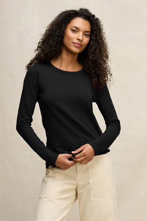 A woman with curly hair wears the LIZZIE TEE by Velvet by Graham & Spencer, a black long-sleeve cotton slub shirt with a classic crew neck, paired with light beige pants against a plain light background.