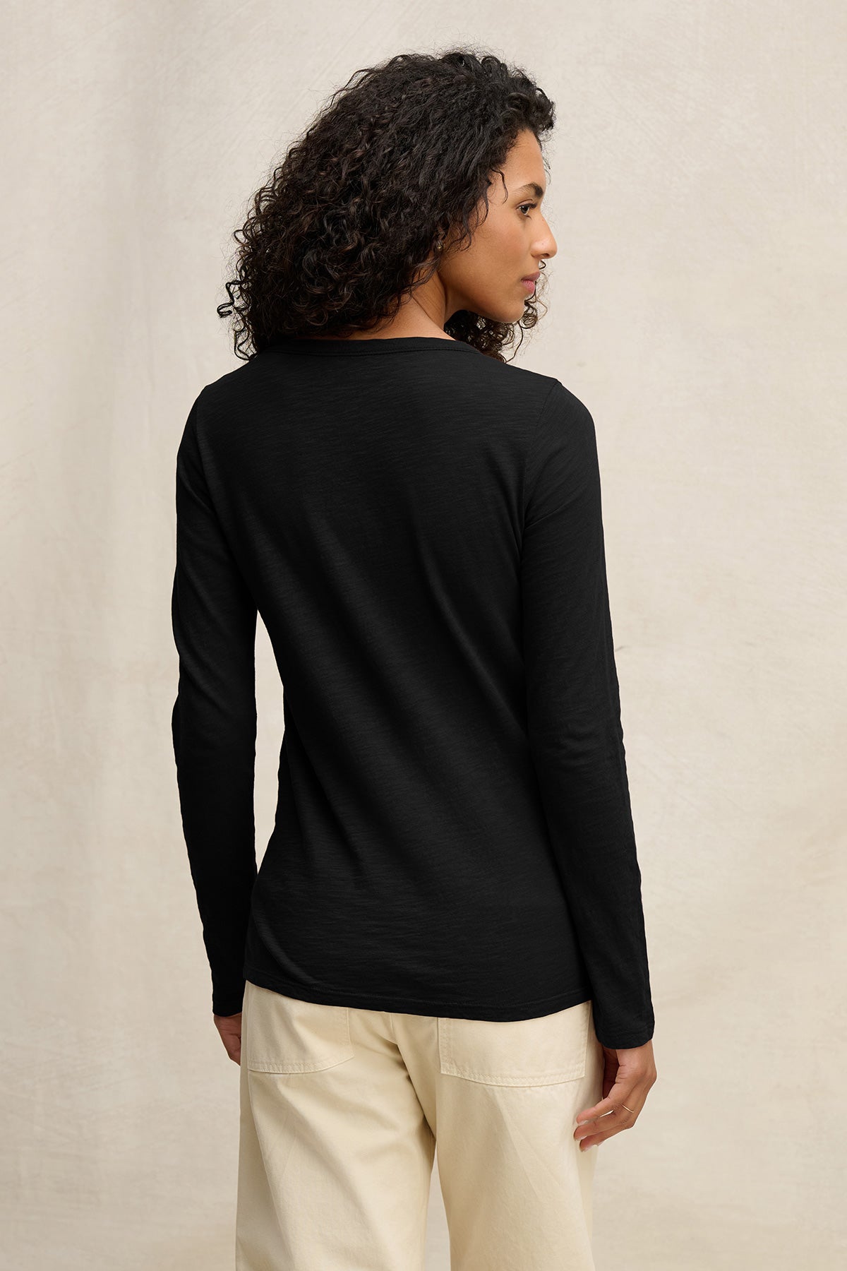 A person with curly hair, dressed in the LIZZIE TEE by Velvet by Graham & Spencer and light-colored pants, stands facing away from the camera against a plain background.-40305805328577