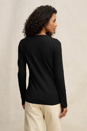 A person with curly hair, dressed in the LIZZIE TEE by Velvet by Graham & Spencer and light-colored pants, stands facing away from the camera against a plain background.