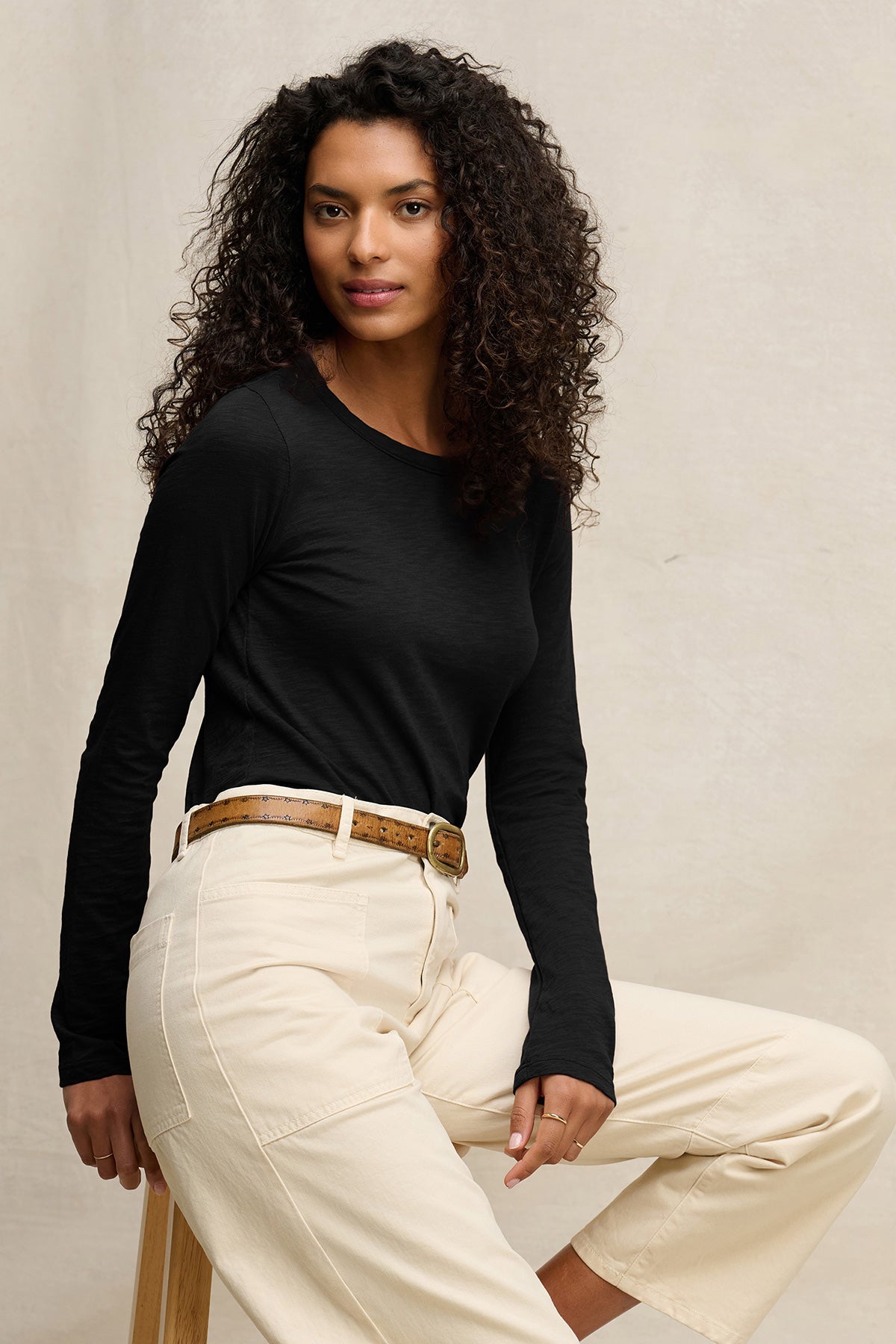 A woman with curly hair wears the LIZZIE TEE by Velvet by Graham & Spencer—a black cotton slub crewneck top—paired with cream pants, sitting on a wooden stool against a neutral background.-40305805230273