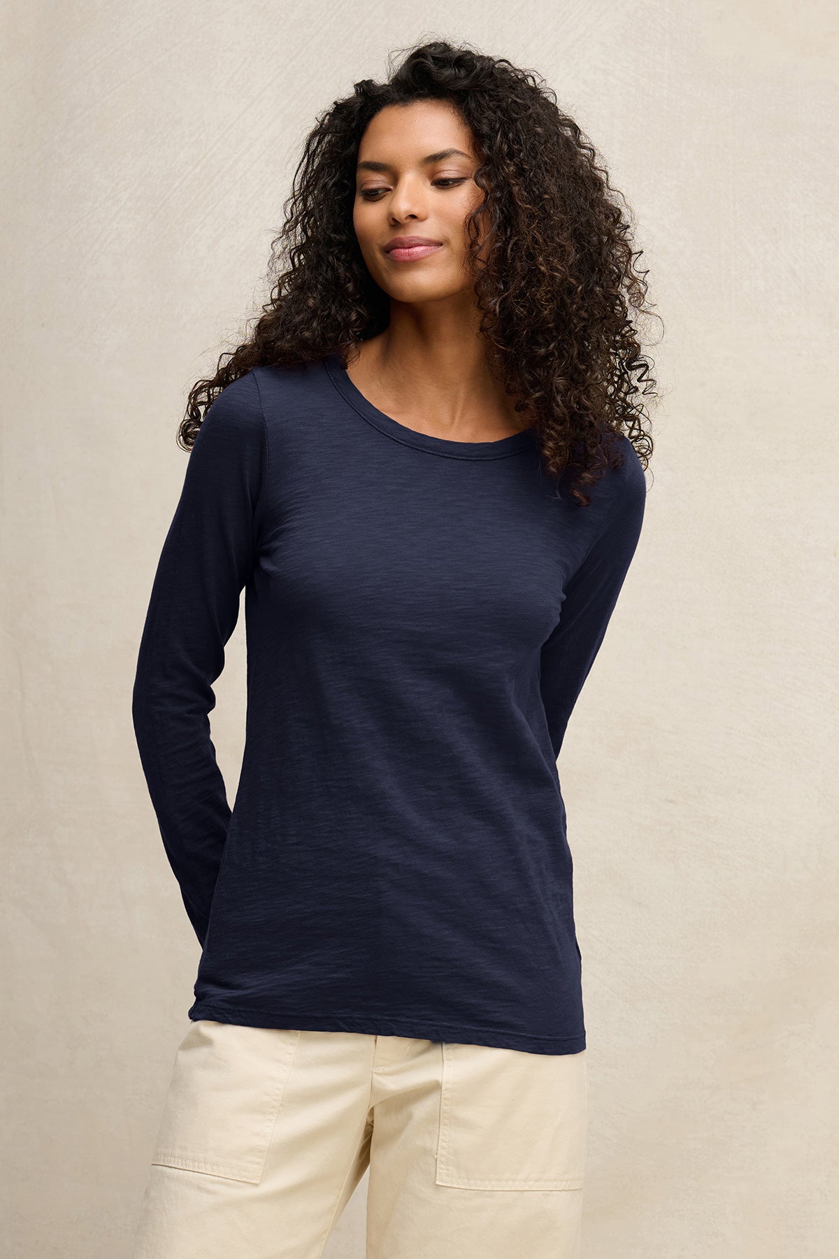 A woman with curly hair wears the LIZZIE TEE by Velvet by Graham & Spencer—a navy cotton slub, long-sleeve shirt—with light-colored pants, standing against a plain, light background.