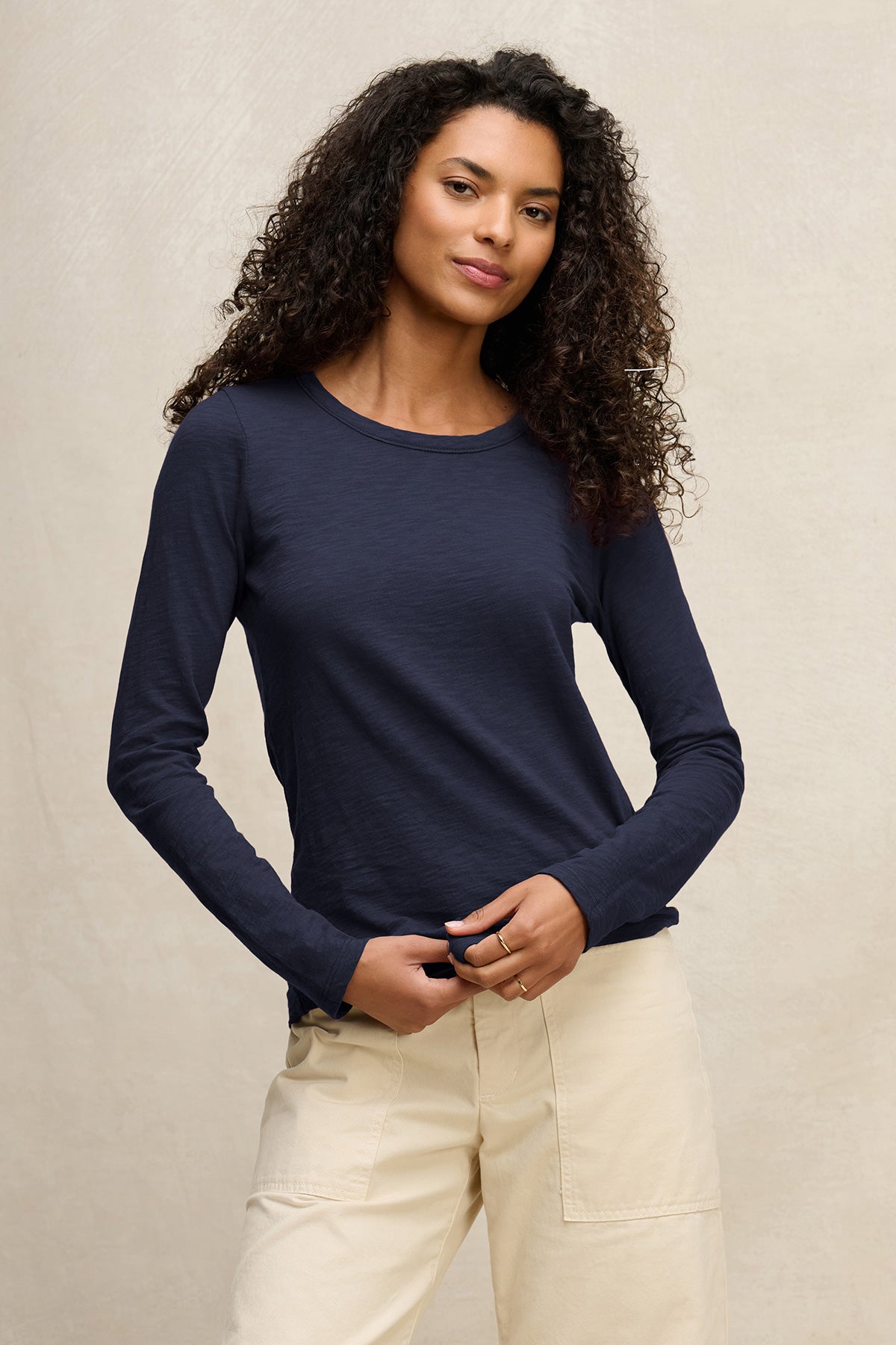 A woman with curly hair wears the LIZZIE TEE by Velvet by Graham & Spencer, a navy cotton slub crewneck, paired with light beige pants against a plain light background.-40306831753409