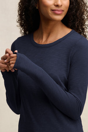 A woman with curly hair wears the LIZZIE TEE by Velvet by Graham & Spencer, a long sleeve dark blue cotton slub top with a crew neckline, holding her hands together near her chest.