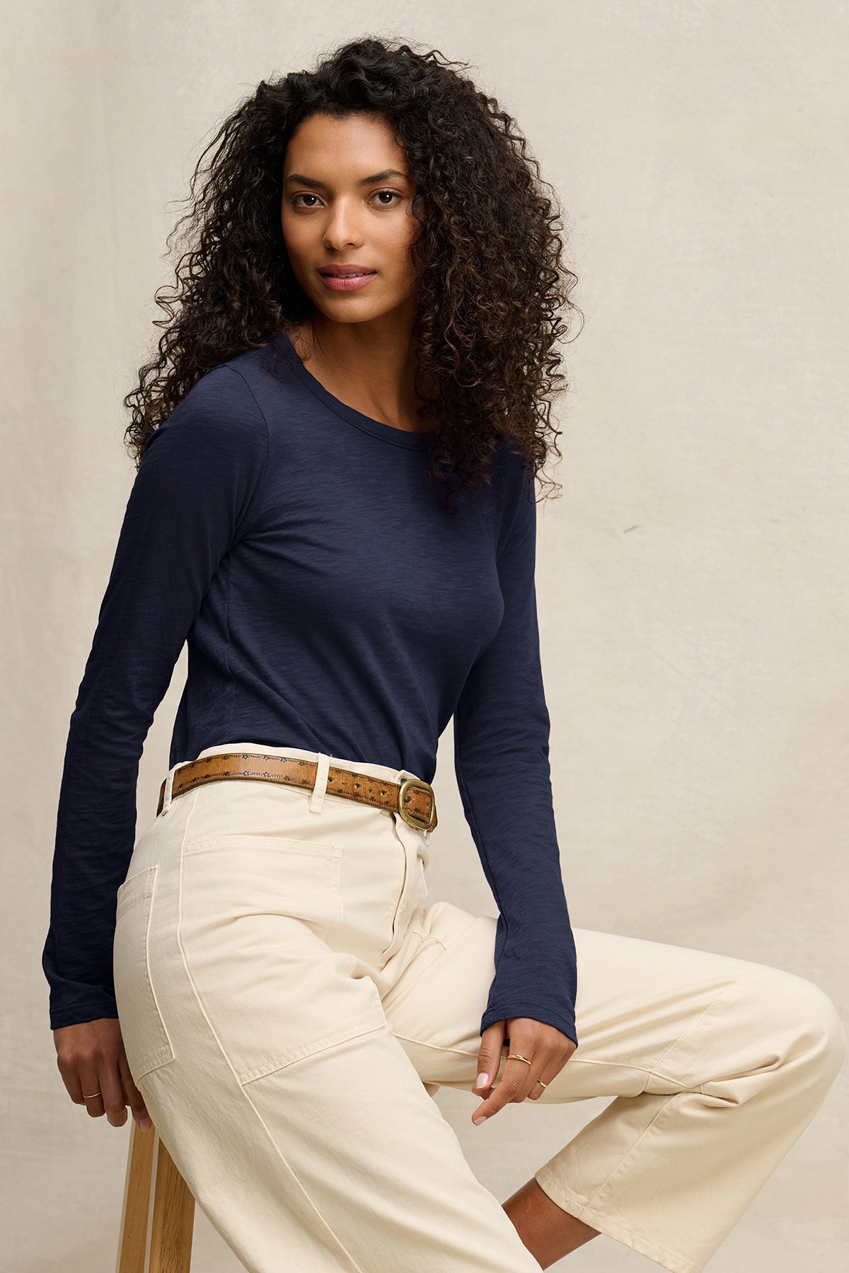A woman with curly hair wears the Velvet by Graham & Spencer LIZZIE TEE in navy, featuring long sleeves and a crew neckline, paired with cream pants as she sits on a stool against a plain light background.