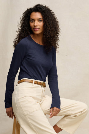 A woman with curly hair wears the Velvet by Graham & Spencer LIZZIE TEE in navy, featuring long sleeves and a crew neckline, paired with cream pants as she sits on a stool against a plain light background.