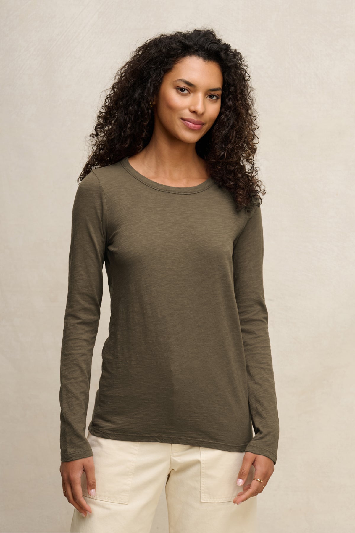 A woman with curly hair wears the LIZZIE TEE by Velvet by Graham & Spencer in olive green with light pants, standing against a neutral background.-40163417522369