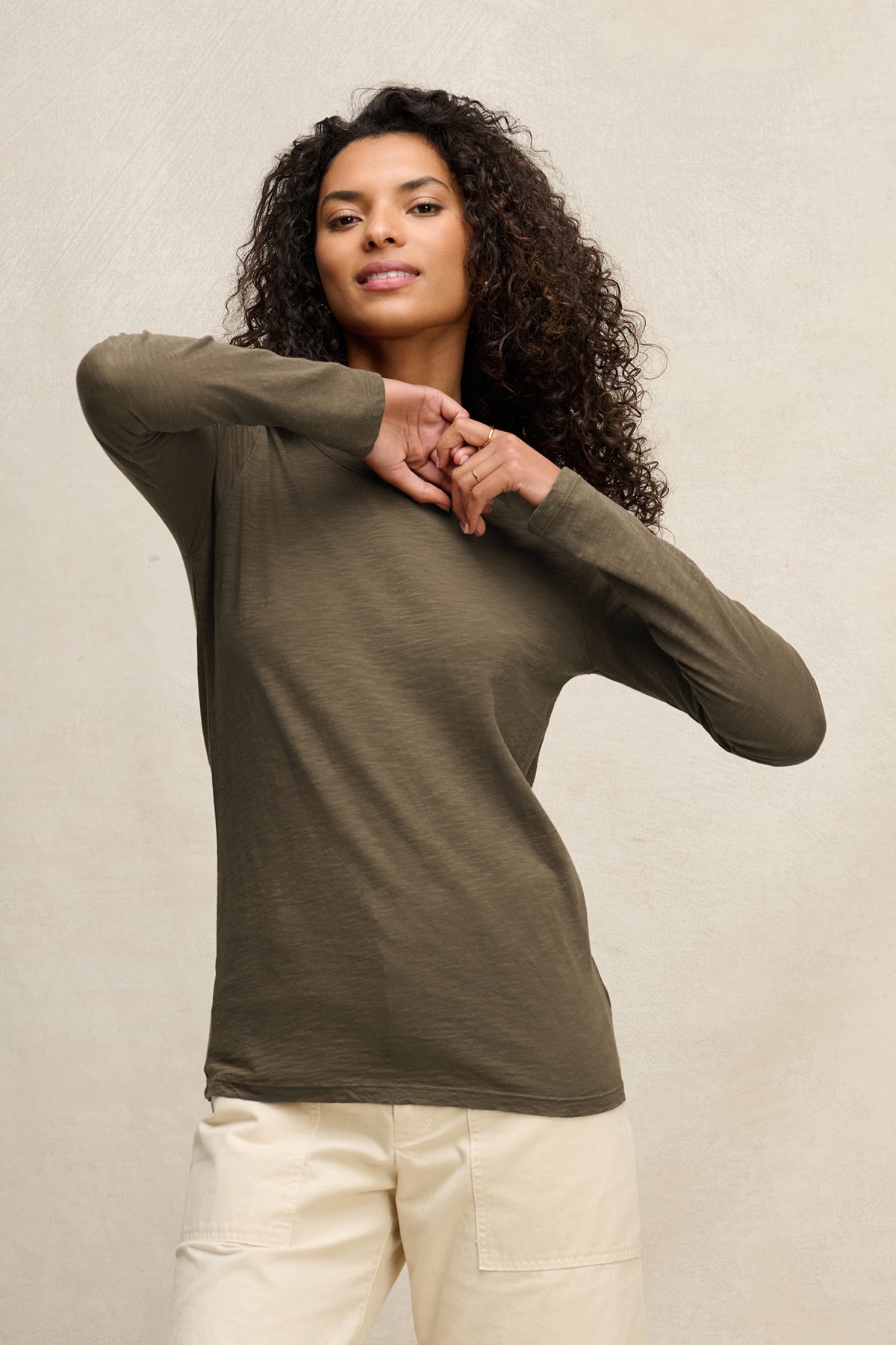 A person models the LIZZIE TEE by Velvet by Graham & Spencer, a long-sleeve olive green shirt, paired with light-colored pants and posing against a neutral background with one arm raised.-40163417456833