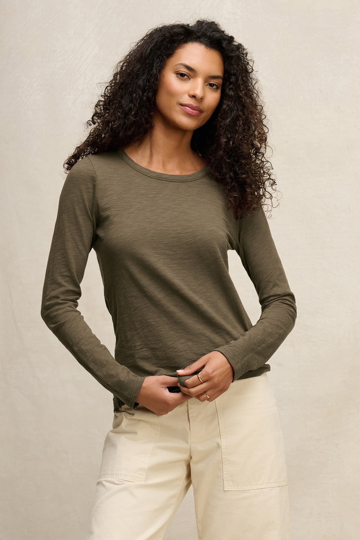A woman with curly hair wears the Velvet by Graham & Spencer LIZZIE TEE, a long-sleeve olive green top, paired with light beige pants, standing against a plain, light-colored background.-40163417424065