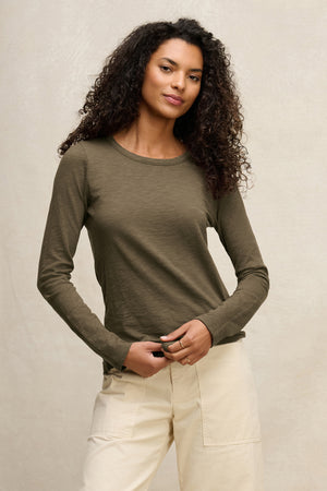 A woman with curly hair wears the Velvet by Graham & Spencer LIZZIE TEE, a long-sleeve olive green top, paired with light beige pants, standing against a plain, light-colored background.