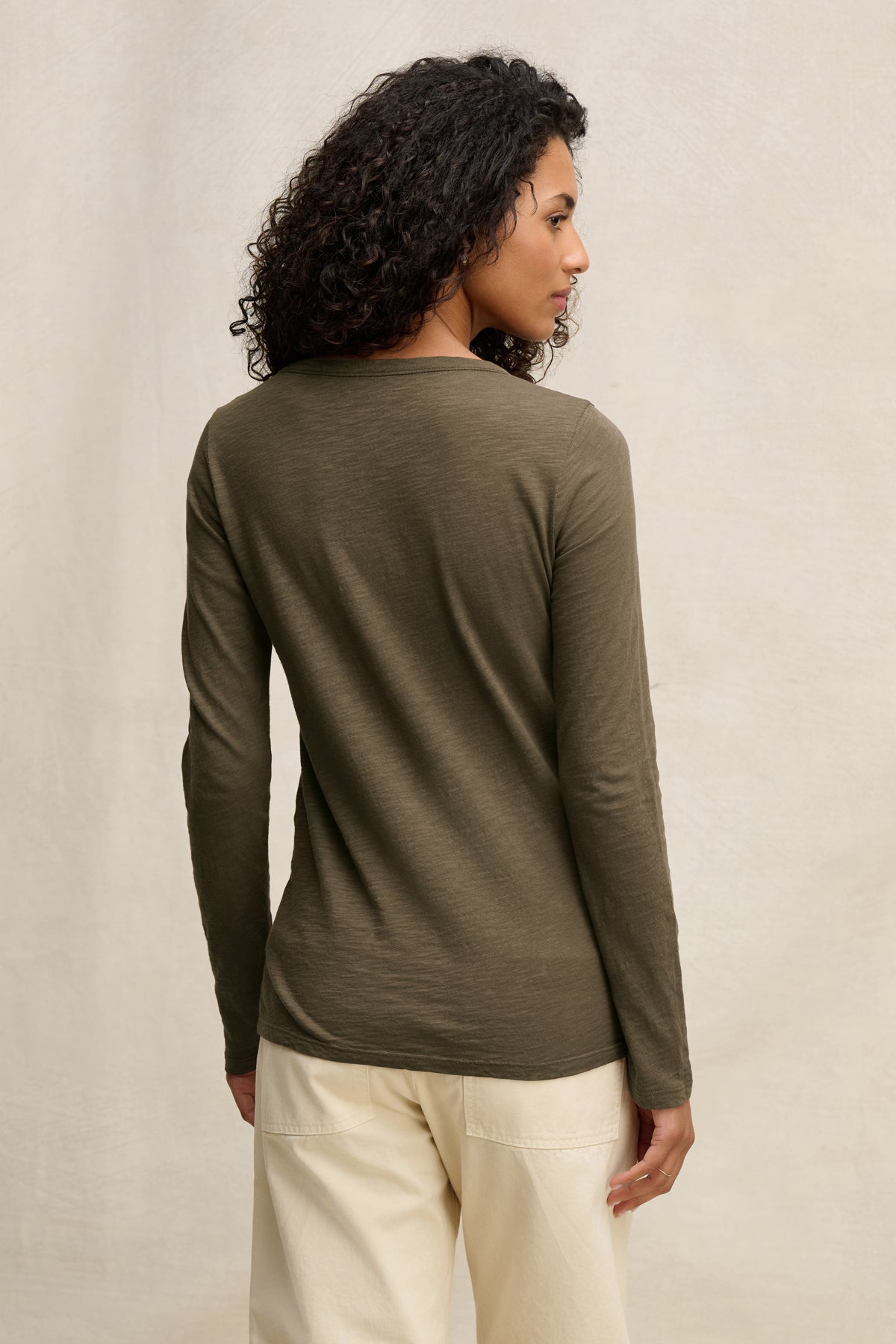 A person with curly hair is shown from the back, wearing the Velvet by Graham & Spencer LIZZIE TEE in olive green with beige pants, posing against a neutral background.