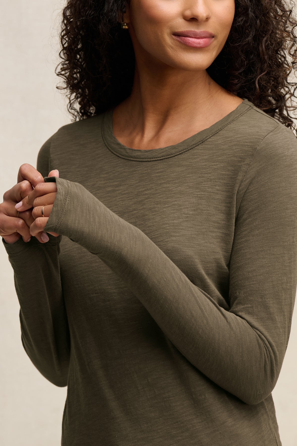 A woman with curly hair wears the Velvet by Graham & Spencer LIZZIE TEE in olive green, standing with her hands clasped and facing slightly to the side.