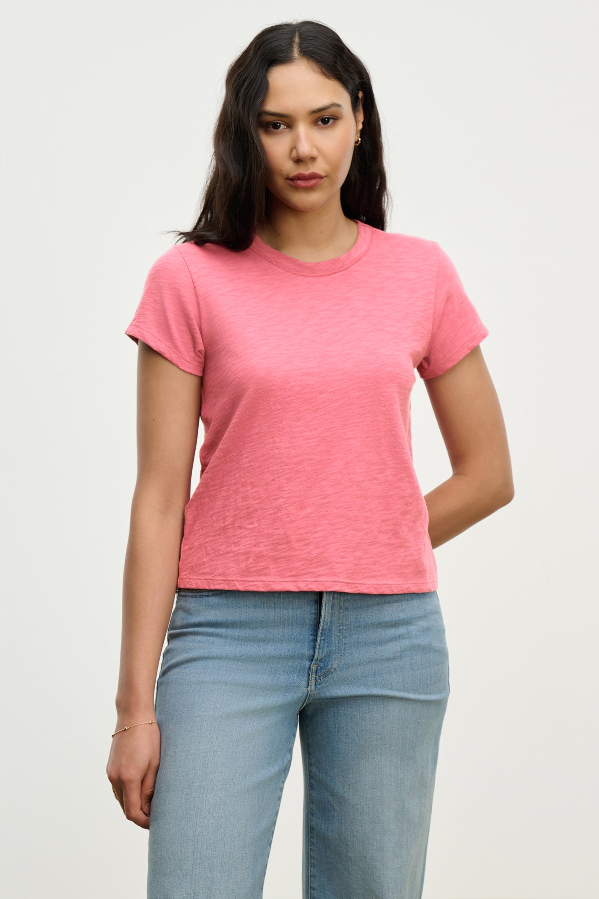   A person with long hair stands against a plain backdrop, wearing Velvet by Graham & Spencer's MARIE TEE in a relaxed boxy fit, paired with light blue jeans. 