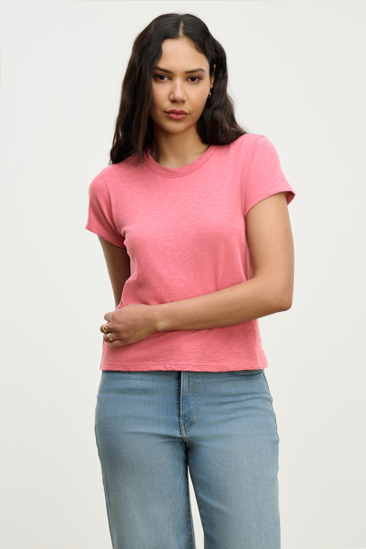   Long-haired individual sports the MARIE TEE from Velvet by Graham & Spencer in soft pink, paired with light blue jeans, highlighting a relaxed boxy fit against a plain backdrop. 