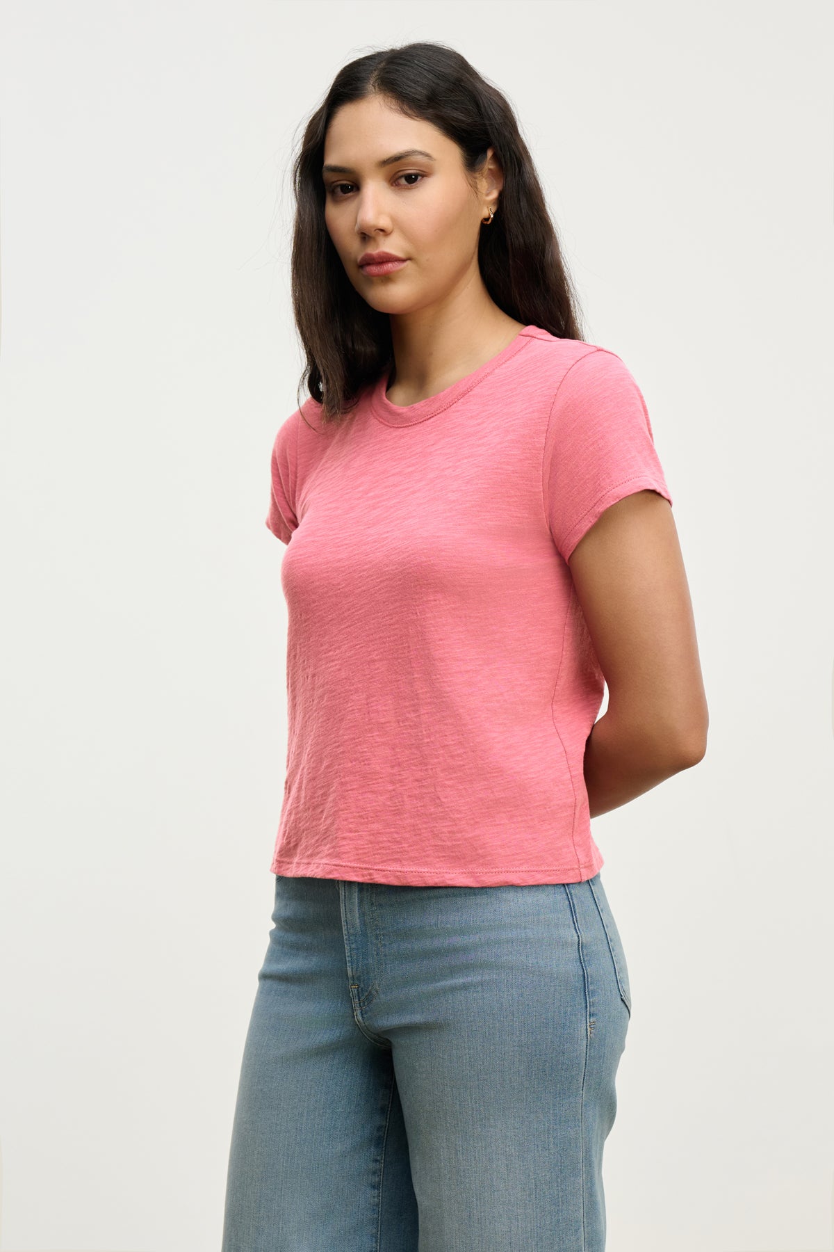   A person with long hair wears the Velvet by Graham & Spencer MARIE TEE, a relaxed boxy fit, along with blue jeans, and stands with their hands behind their back against a plain background. 